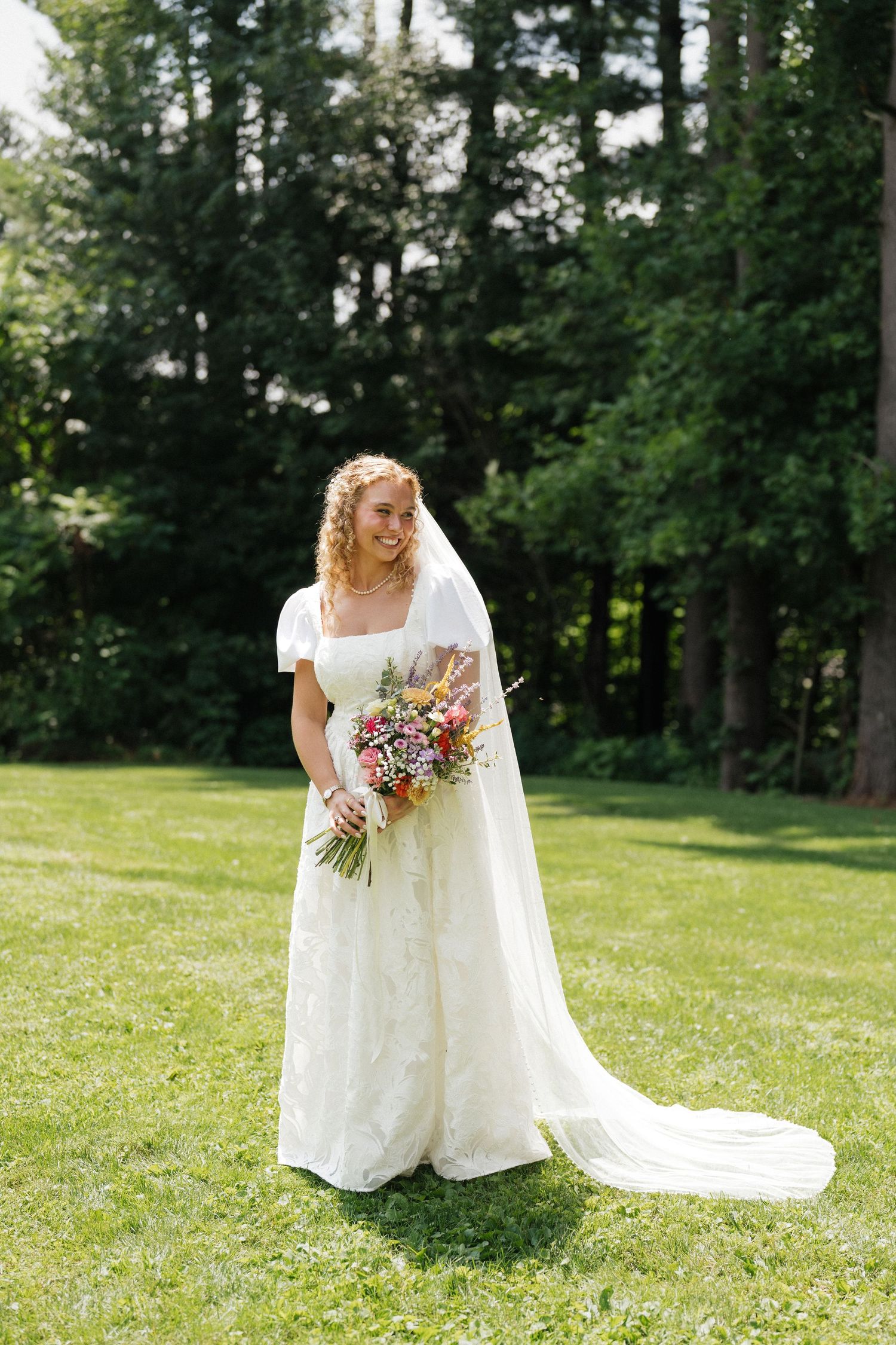 Smiling bride in white lace gown with veil holds colorful wildflower bouquet on green lawn with tall trees behind her.