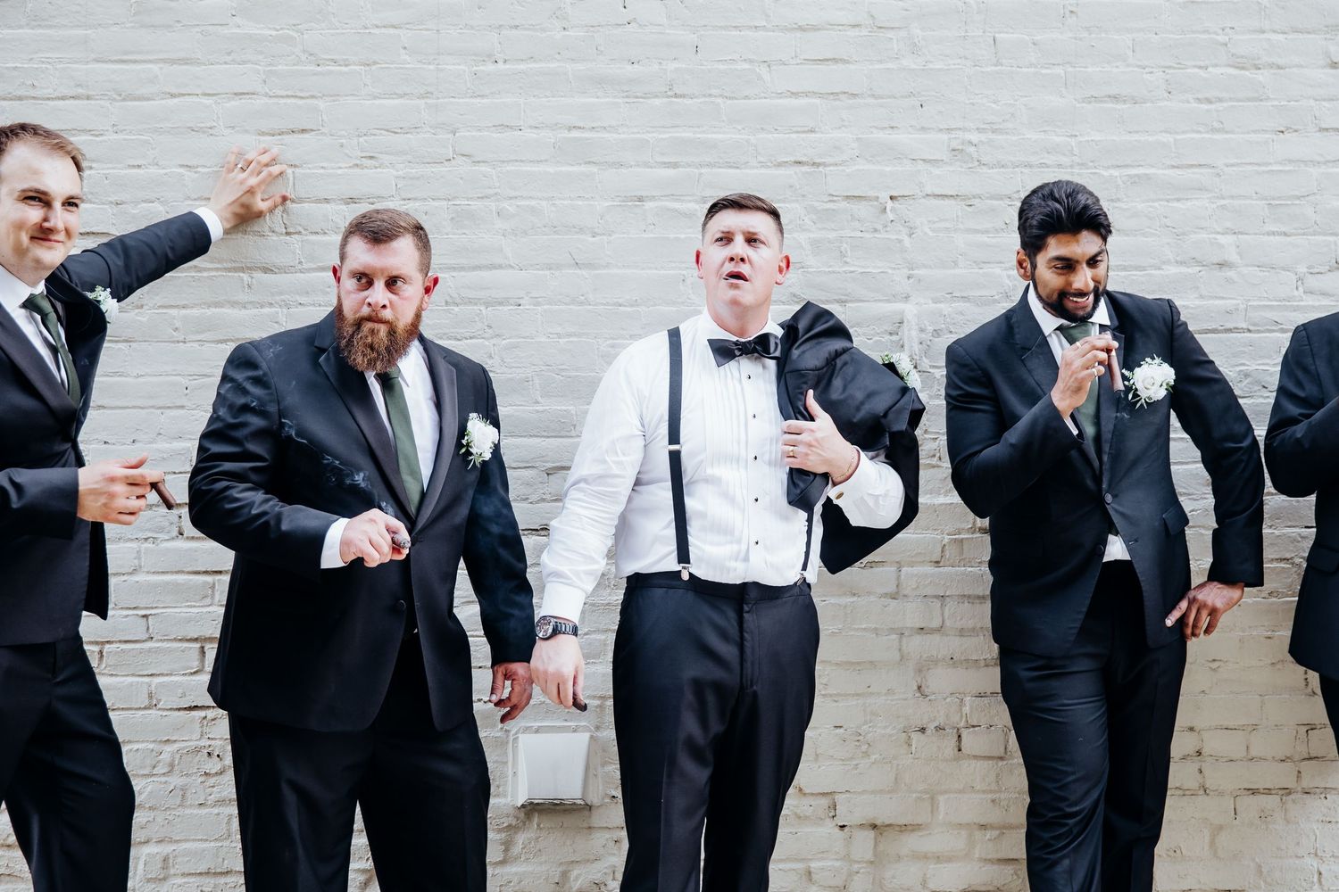 Groomsmen in black suits perform animated poses against a white brick wall.