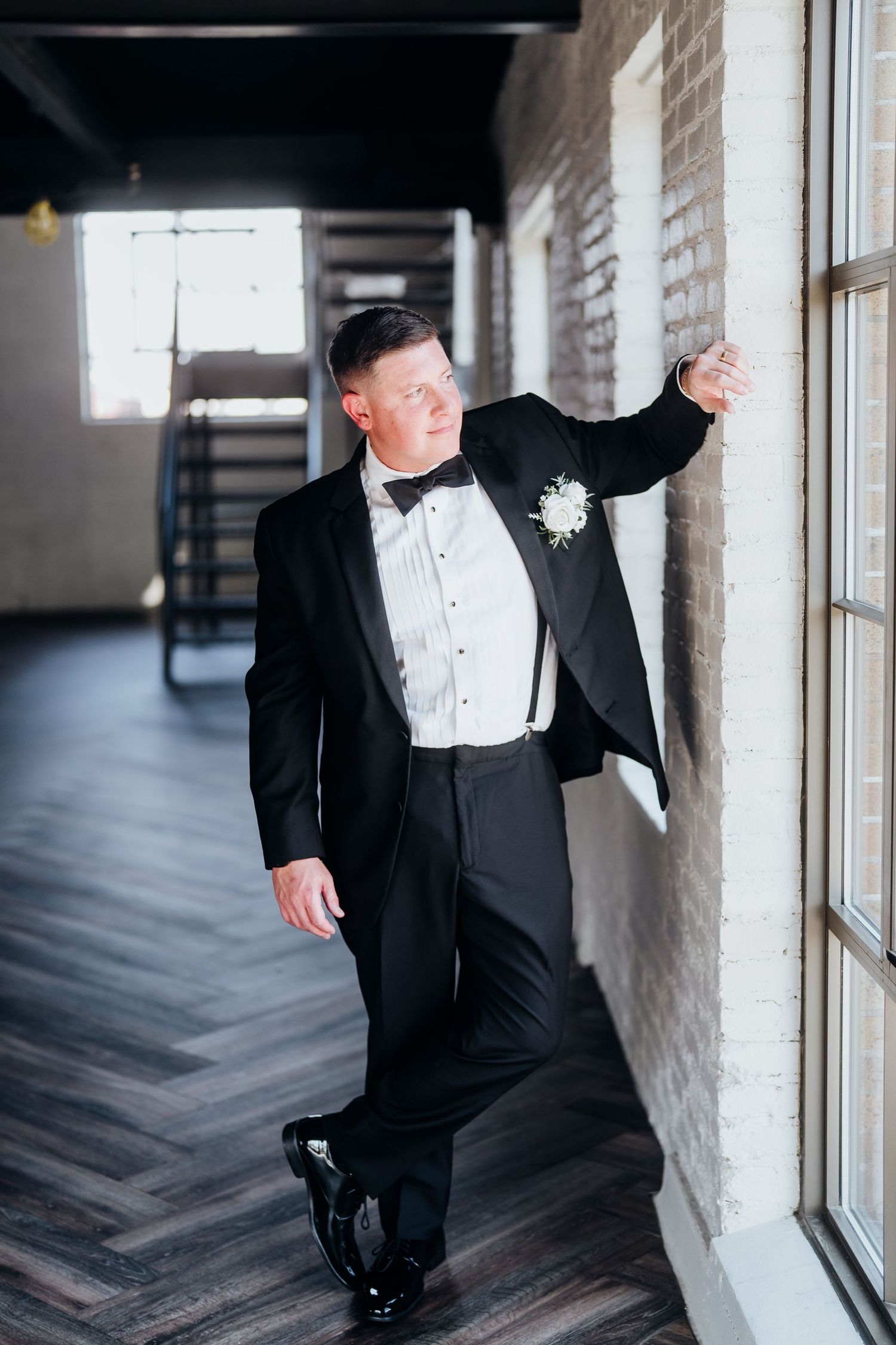 A groom in a classic black tuxedo poses in a rustic brick interior with large windows.