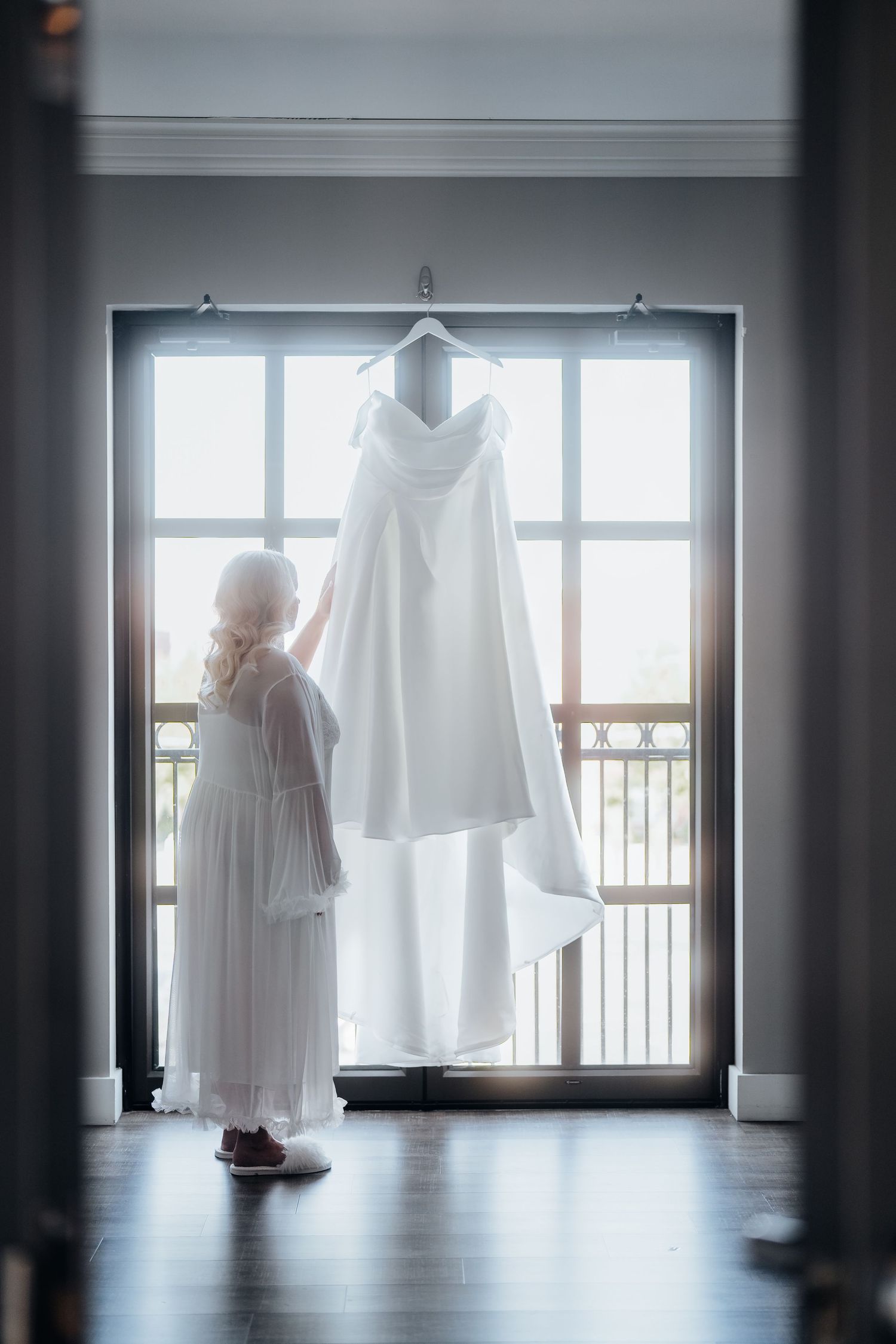 Elegant white wedding dress hanging in window silhouette with natural backlight.