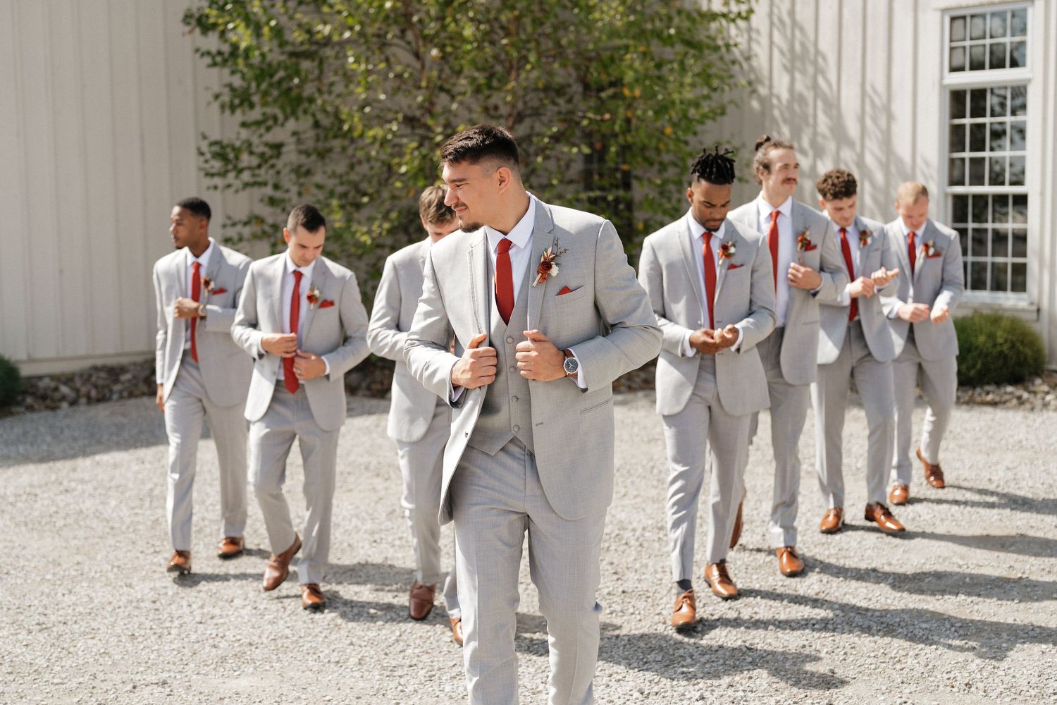 Wedding party members in light gray suits and red ties stand outside a white building.