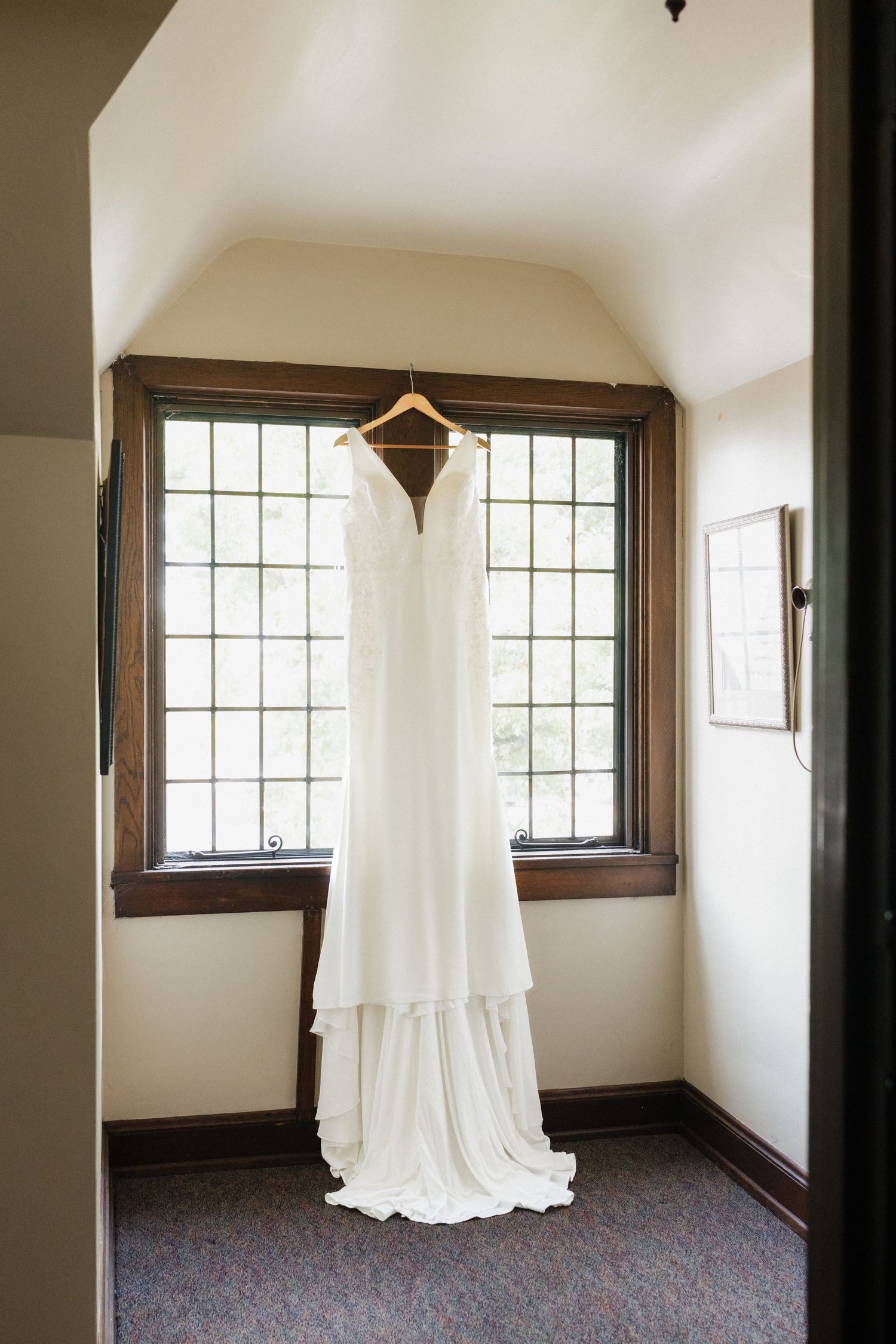 A white wedding dress hangs elegantly in a room with large windows and dark wood trim.