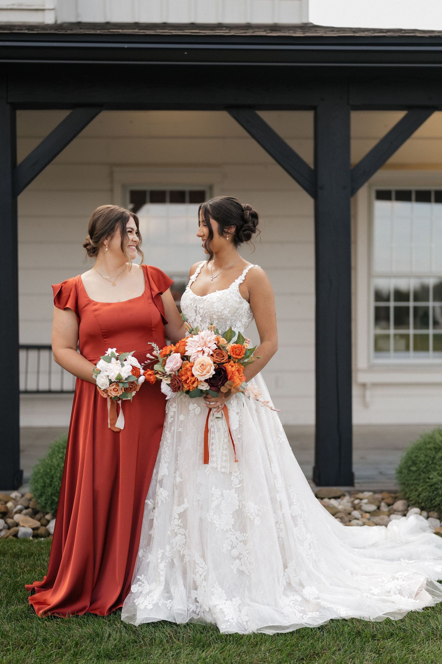 A bride in white and bridesmaid in rust-colored dresses sit together holding bouquets outdoors.