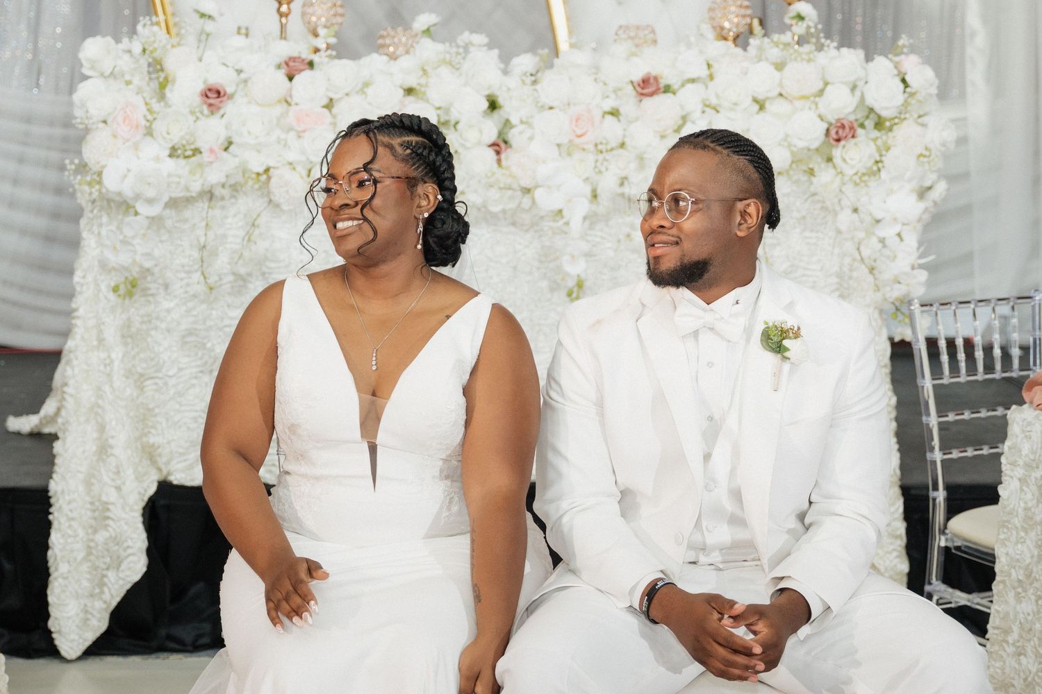 Wedding couple seated together in front of elegant white floral backdrop.