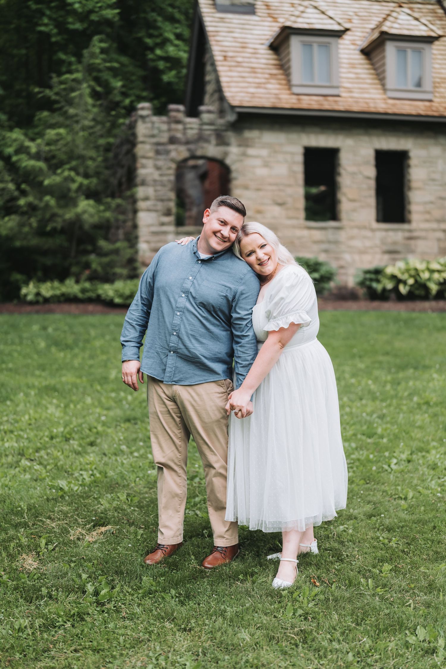 A couple poses together on a lawn in front of a stone cottage.