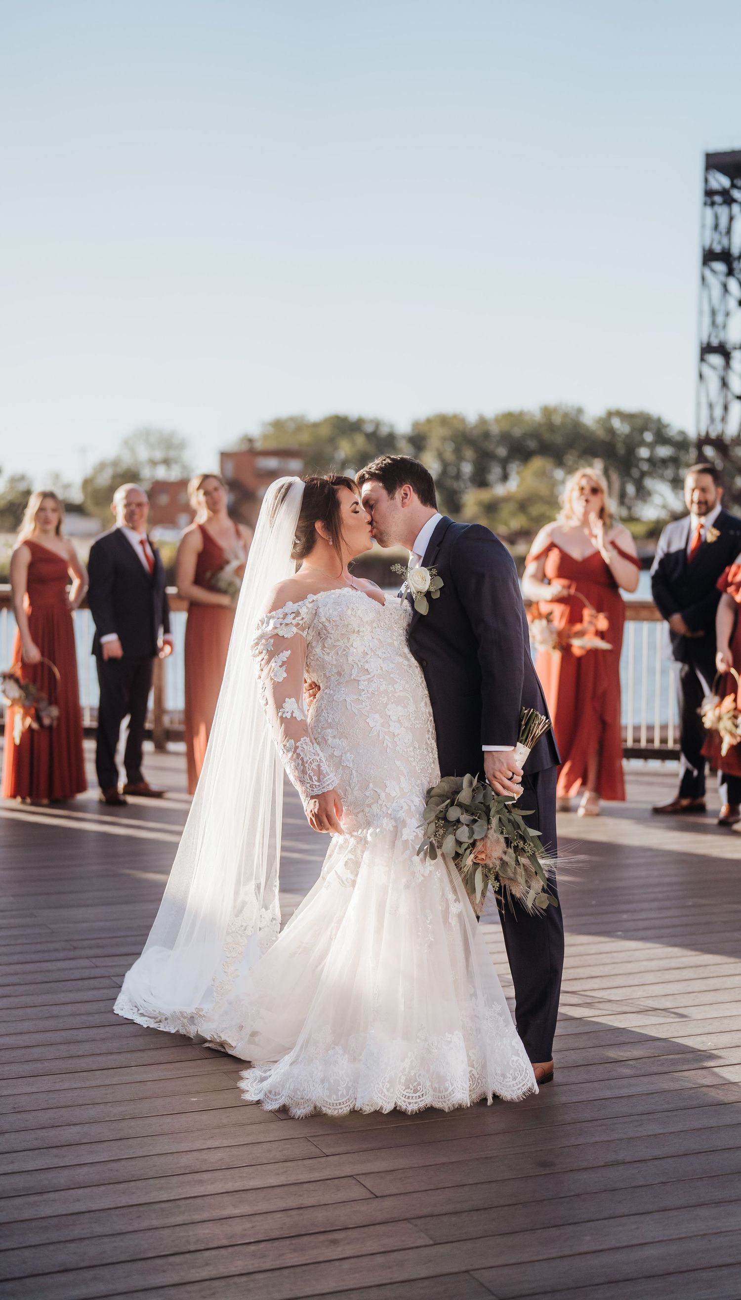 Wedding ceremony on a wooden deck with bridesmaids in rust-colored dresses looking on.