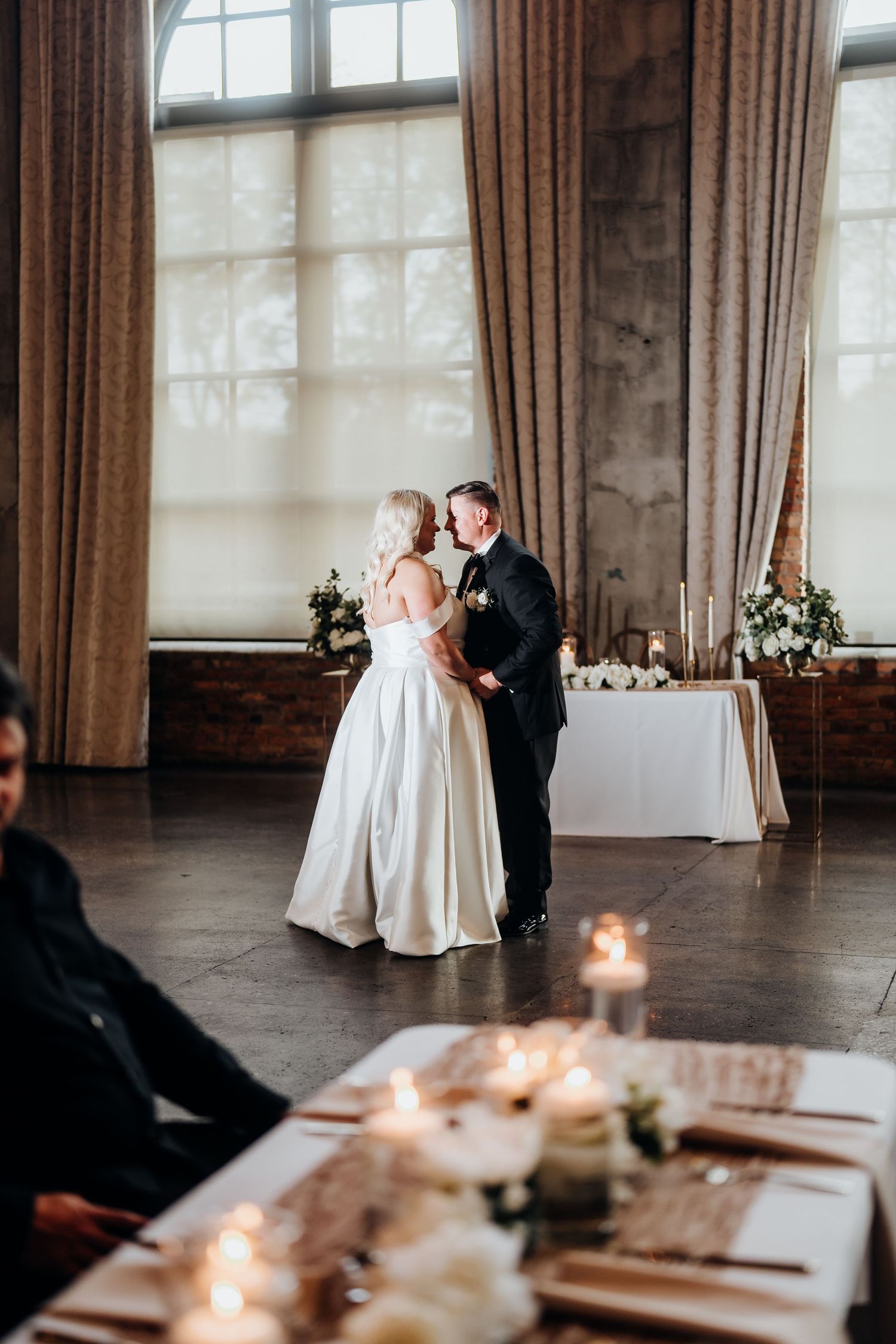 Romantic first dance in an industrial-chic venue with candlelit tables in the foreground.