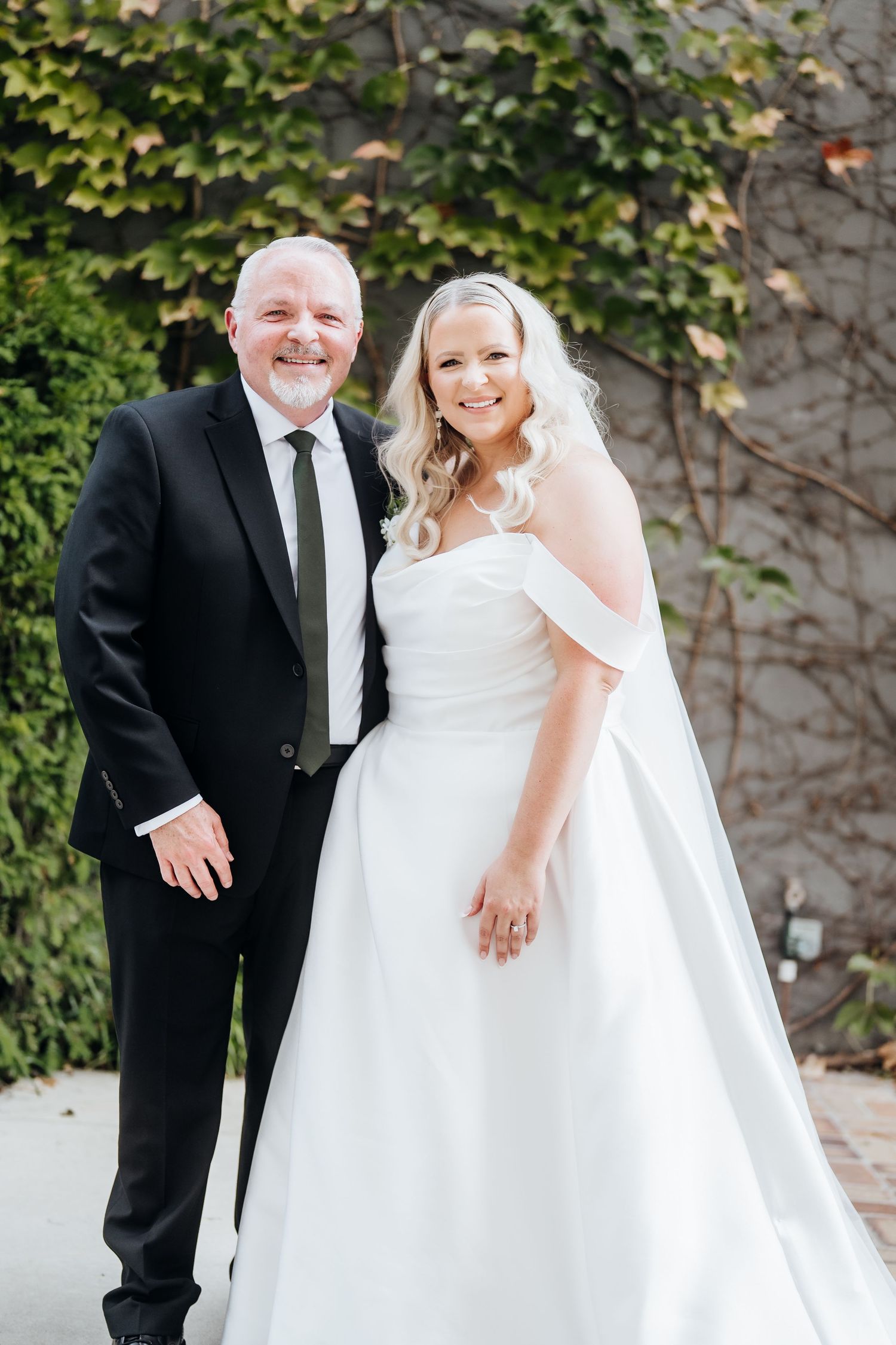 Bride in elegant white ball gown poses with companion against ivy-covered wall.