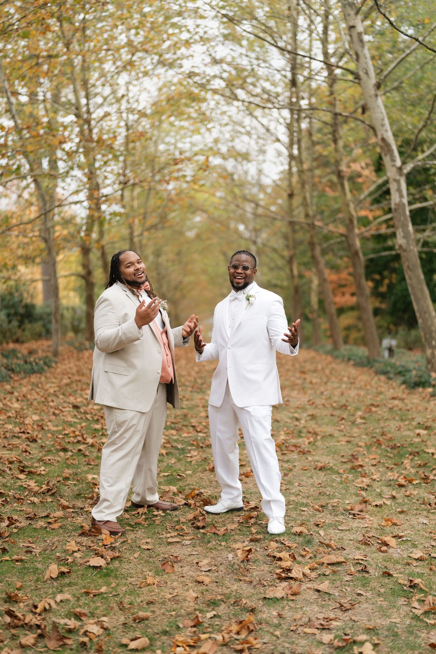 Two people in white outfits share a joyful moment on fallen autumn leaves in a wooded setting.