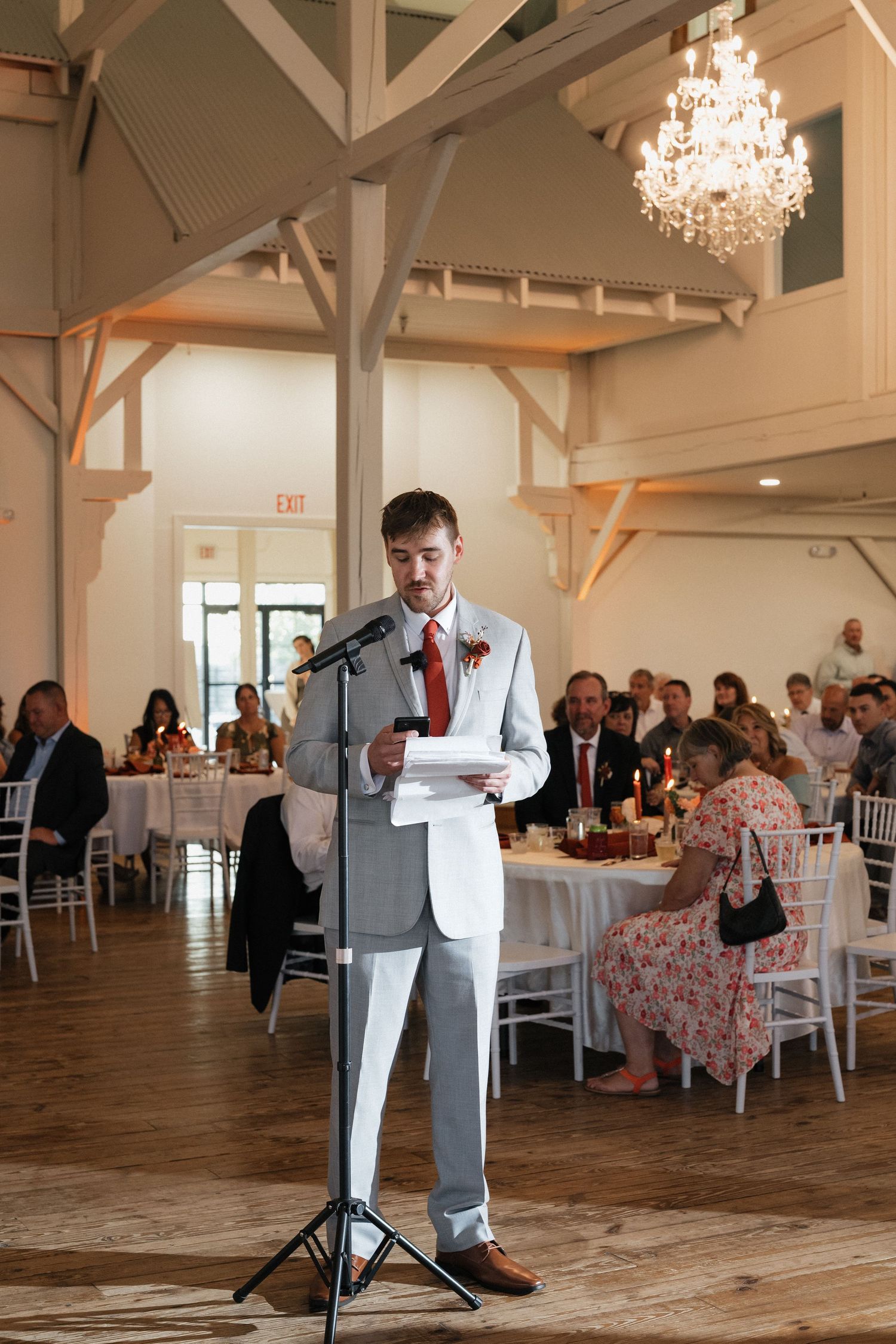 Wedding speech being delivered in an elegant white barn venue with exposed beams and crystal chandeliers.