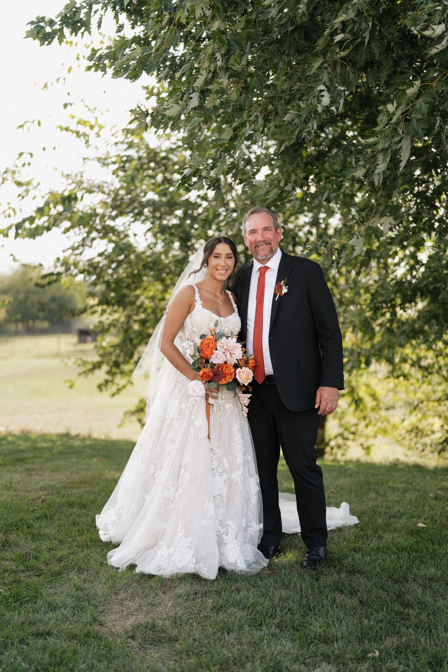 Wedding portrait of couple with orange bouquet under tree branches.