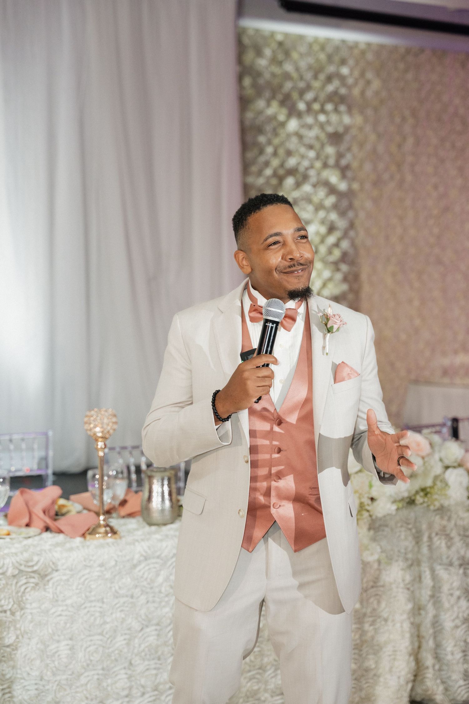 Person in cream suit and coral vest speaks into microphone at wedding reception.
