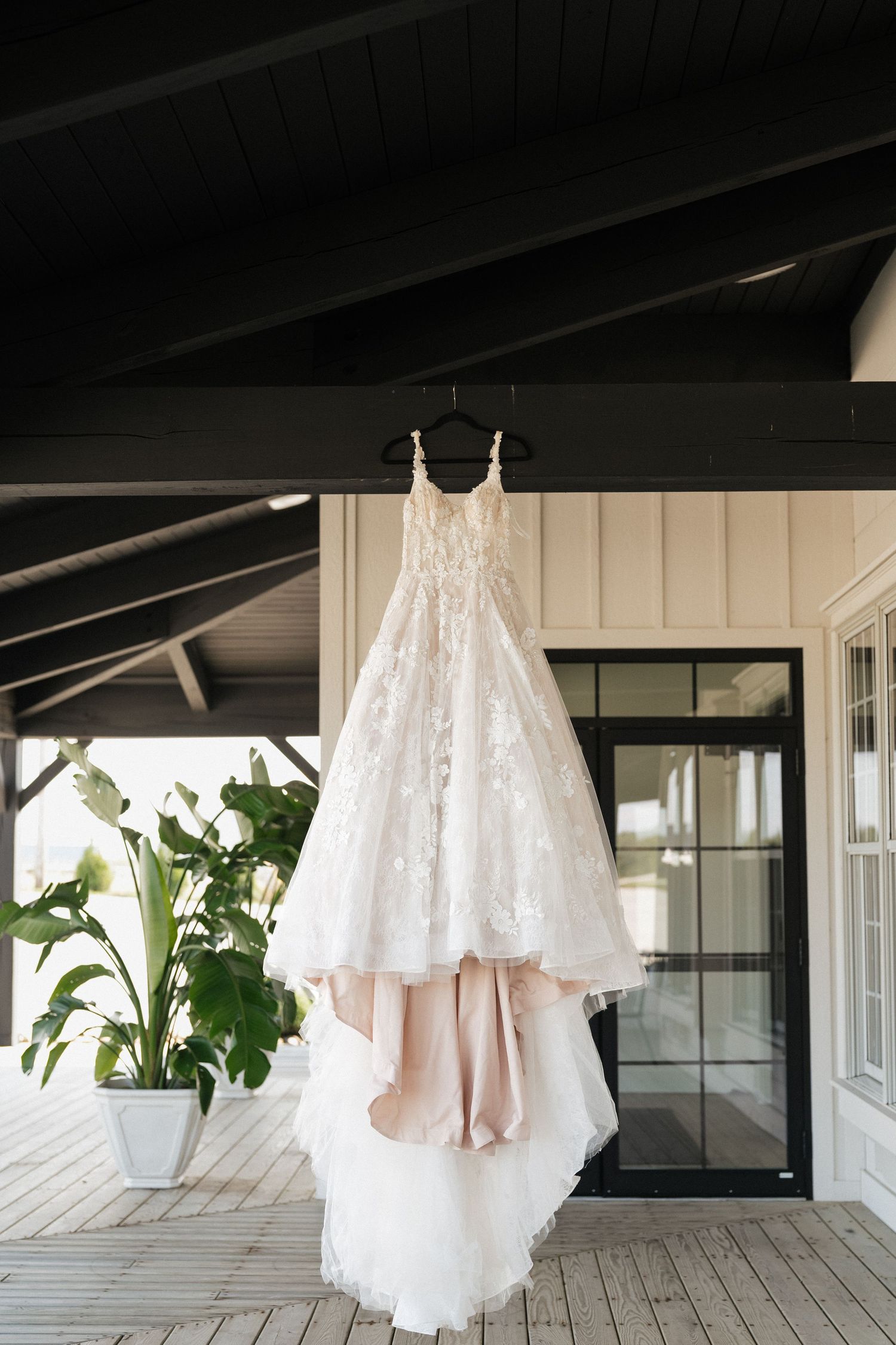 White wedding dresses hang elegantly against a light-colored wall on a covered porch with green plants visible.