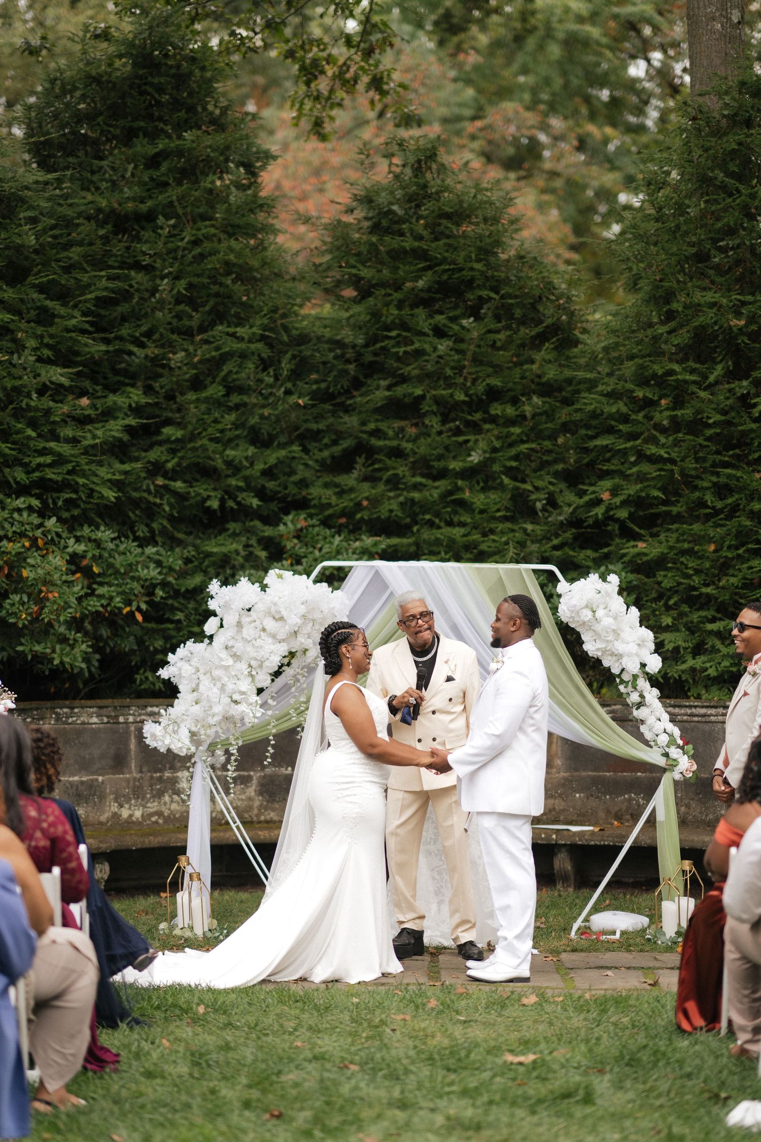 Wedding ceremony takes place under a white floral arch with evergreen trees in the background.