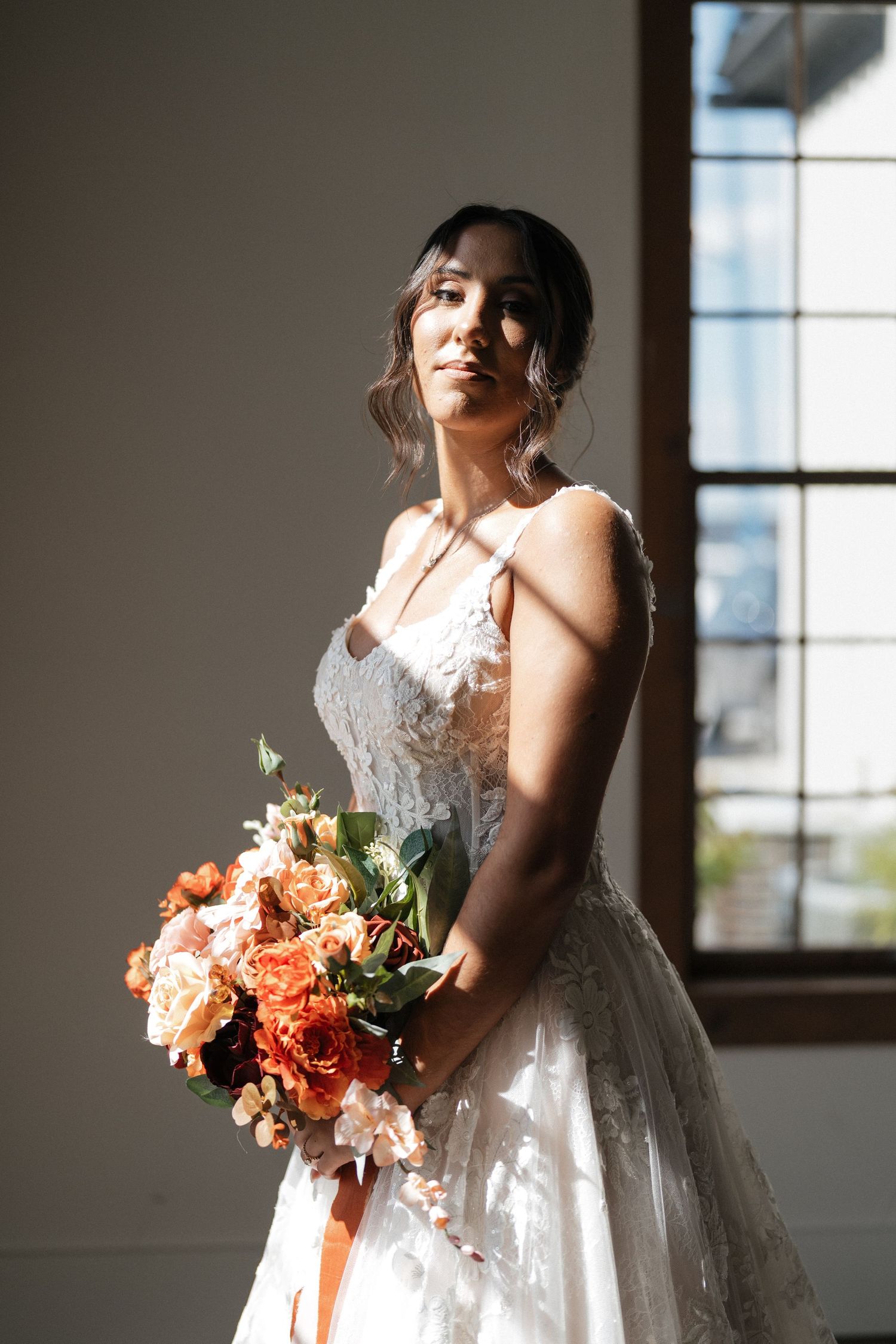 Bride in elegant white gown holds a coral-colored bouquet in natural window light.
