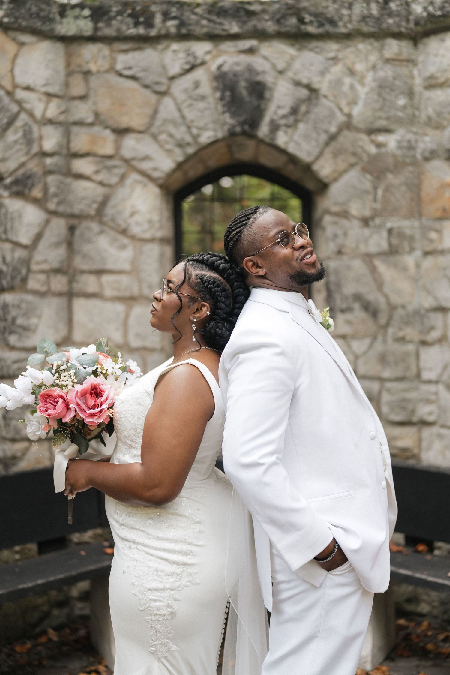 Newlyweds pose playfully back-to-back against a rustic stone wall backdrop.