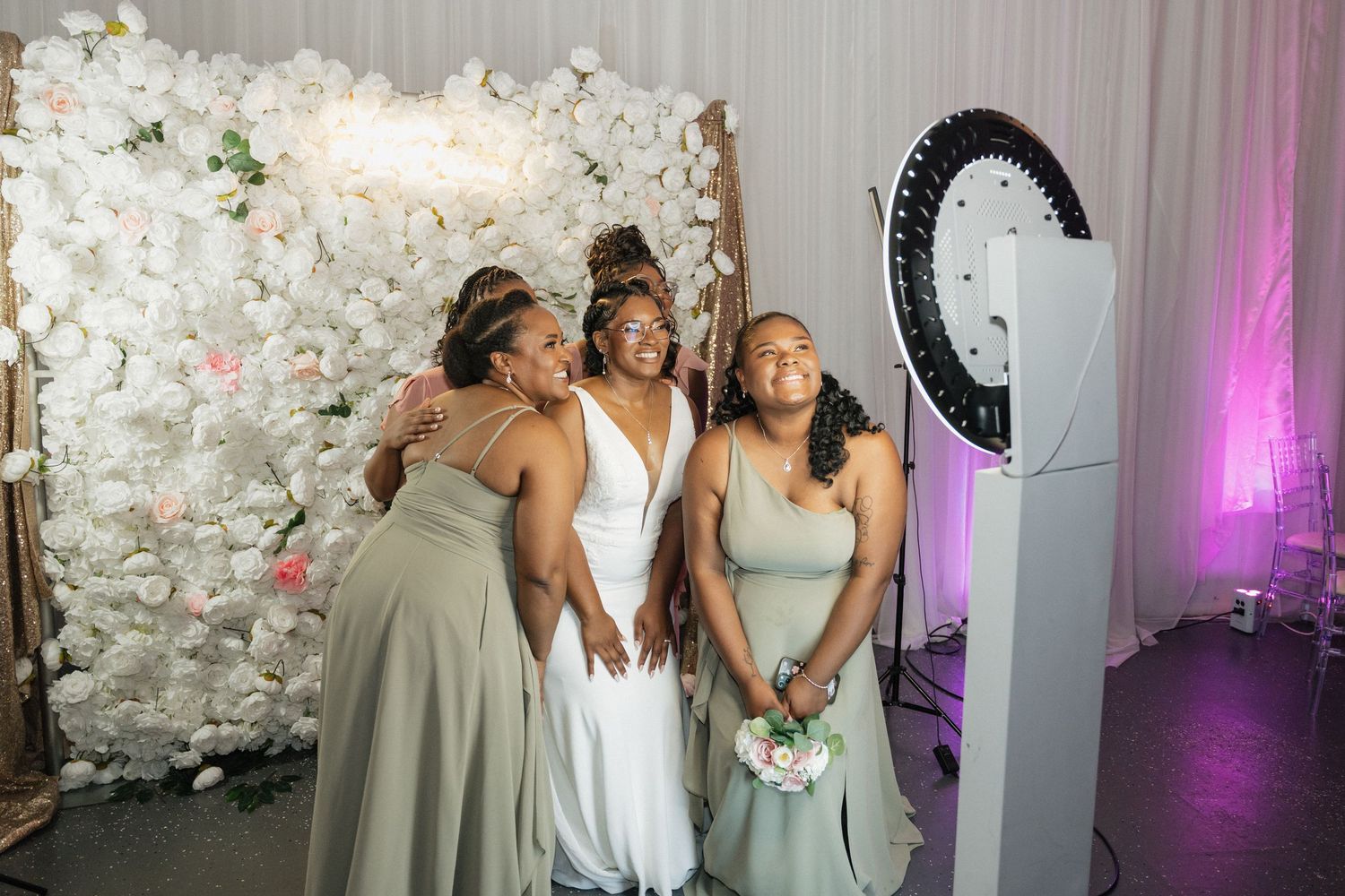 Wedding party poses together at a photo booth with white floral backdrop and ring light.