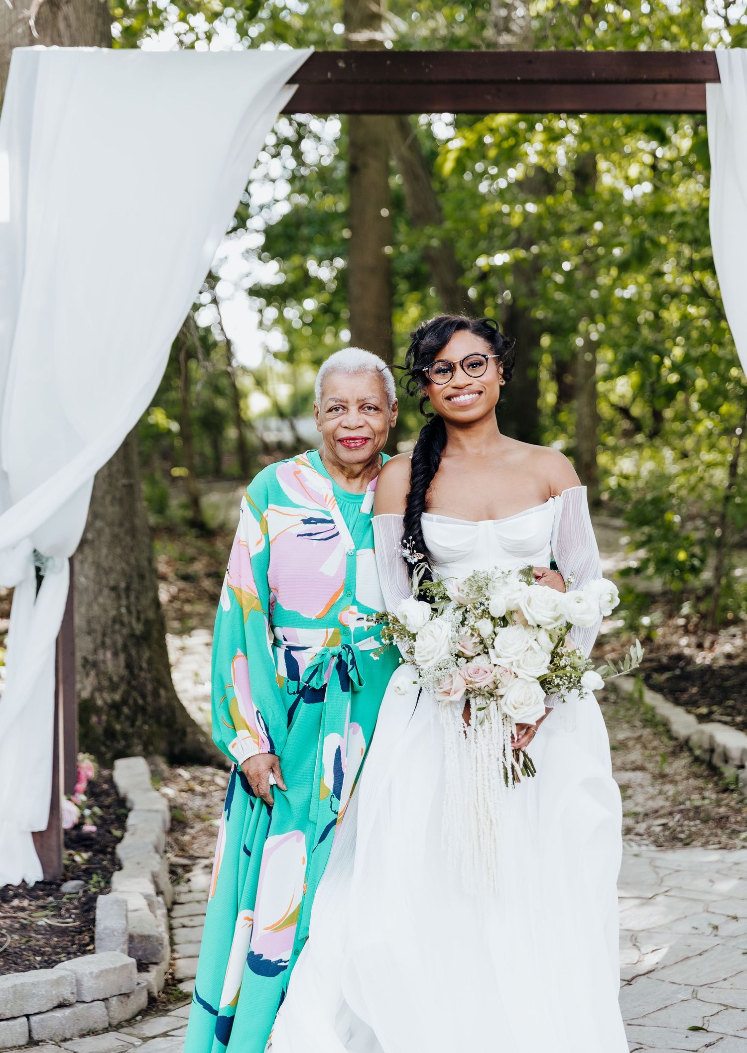 A bride in white gown stands with family member wearing vibrant turquoise patterned outfit under wedding arch.