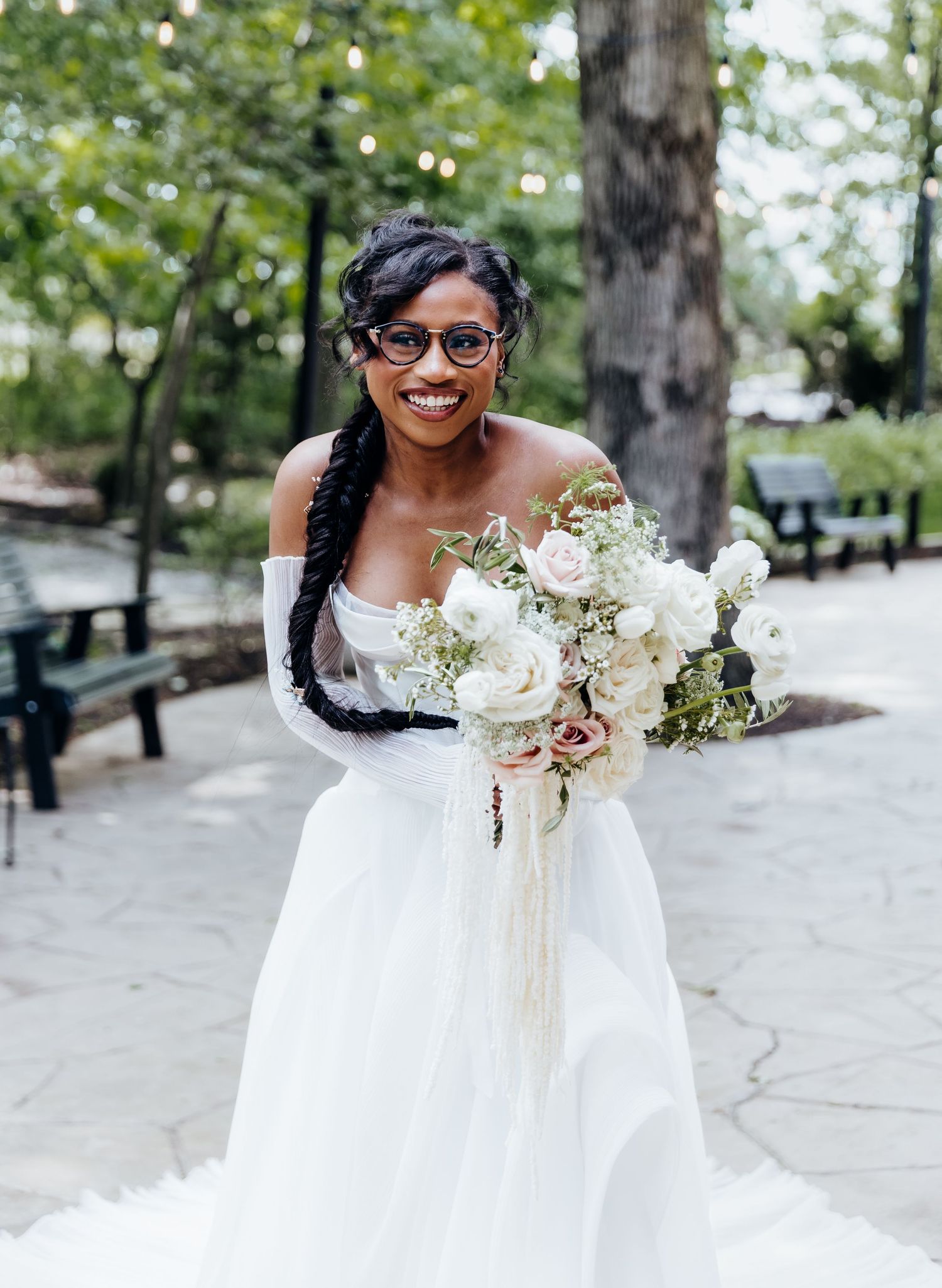 Bride in flowing white dress holds romantic bouquet of roses and greenery in outdoor setting.