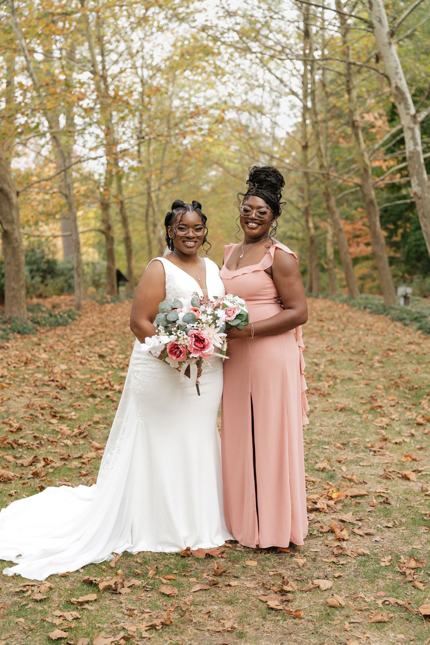 A bride and bridesmaid pose together holding a bouquet in a fall forest setting.