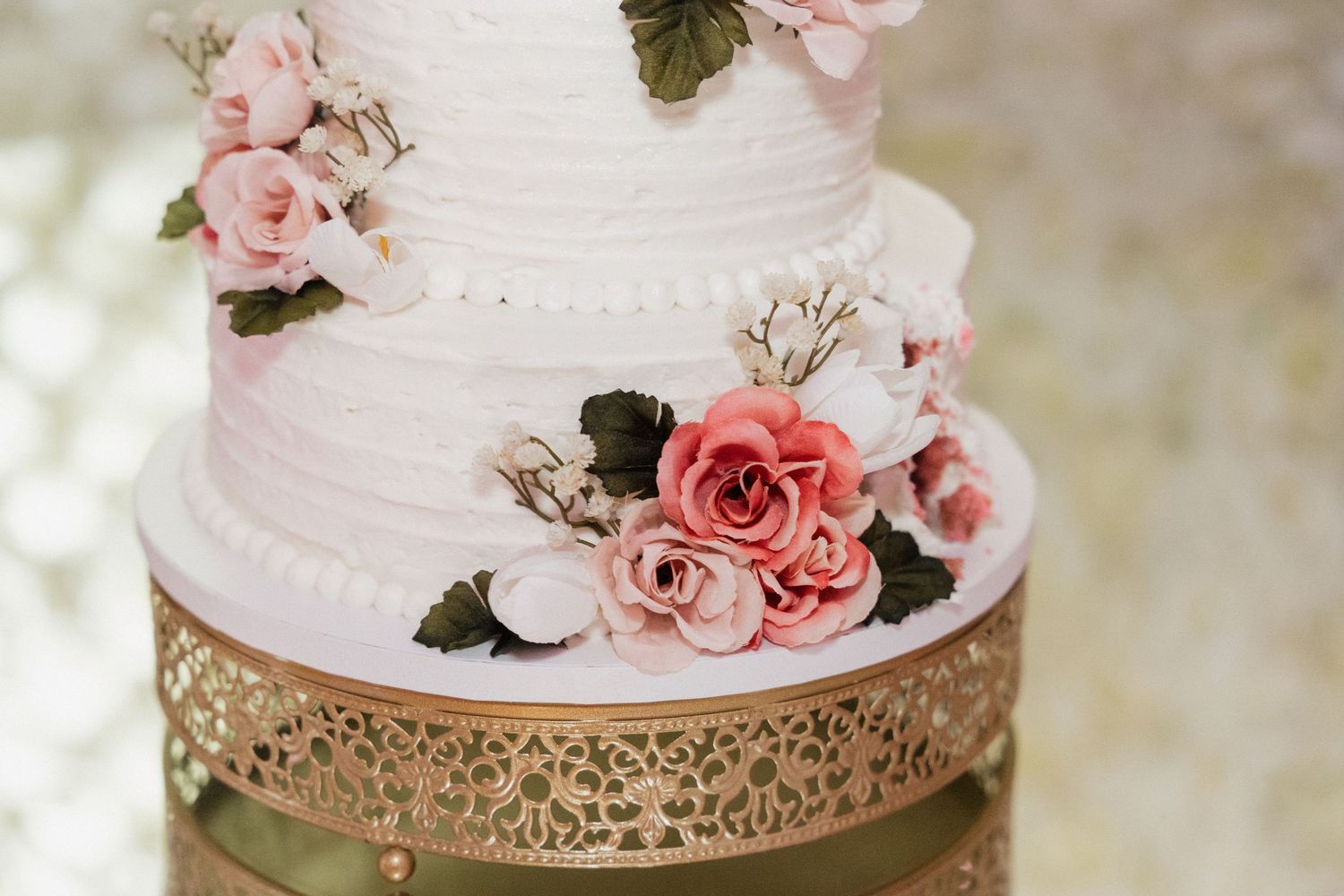 Elegant white wedding cake decorated with pink roses on gold filigree stand.