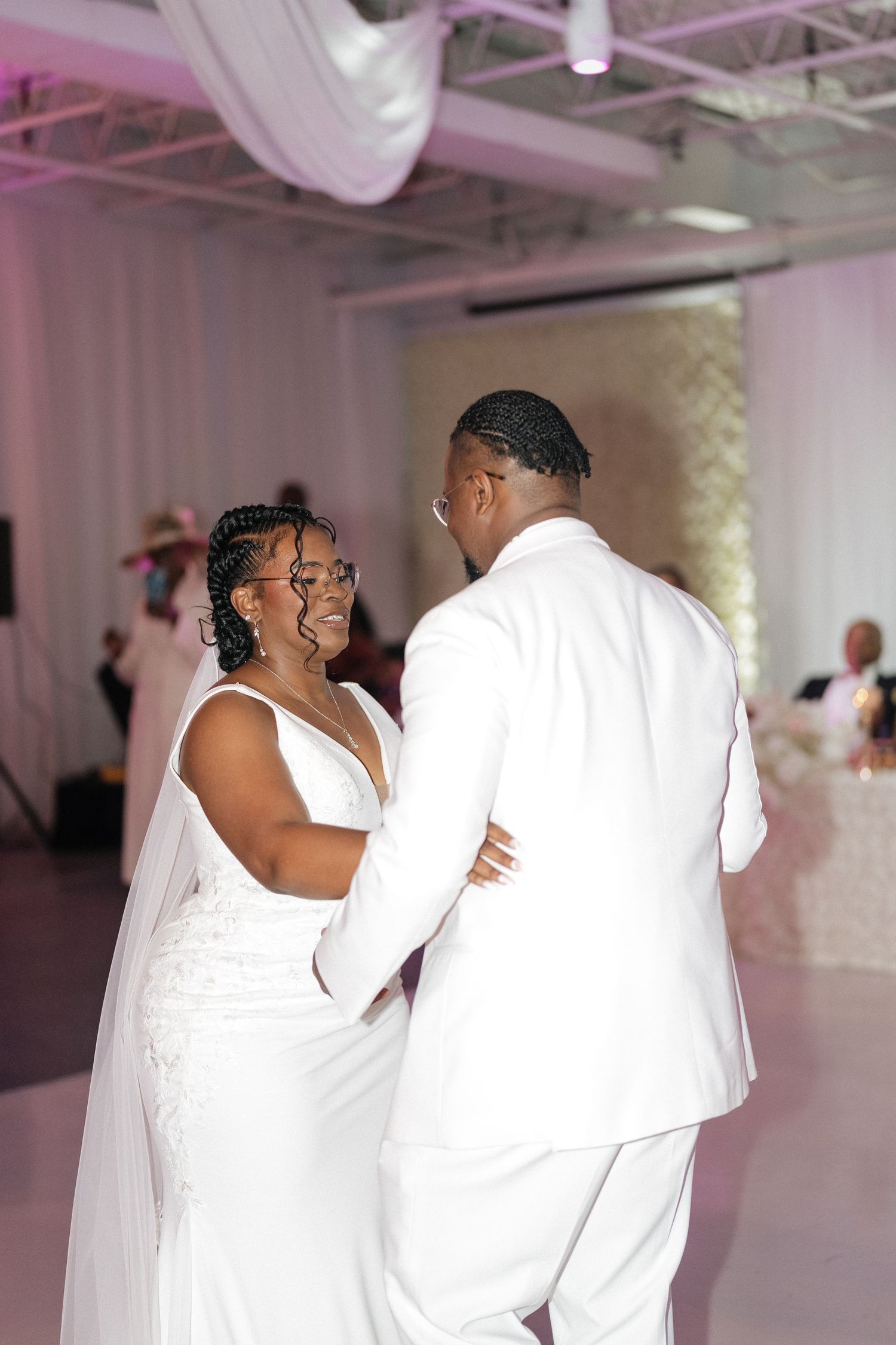 Wedding couple shares intimate dance moments in an elegant white reception venue with pink lighting.