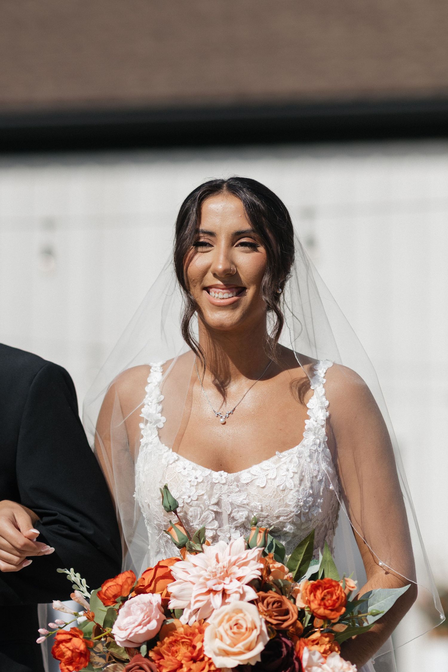 Bride in white lace dress holds vibrant coral and peach rose bouquet.