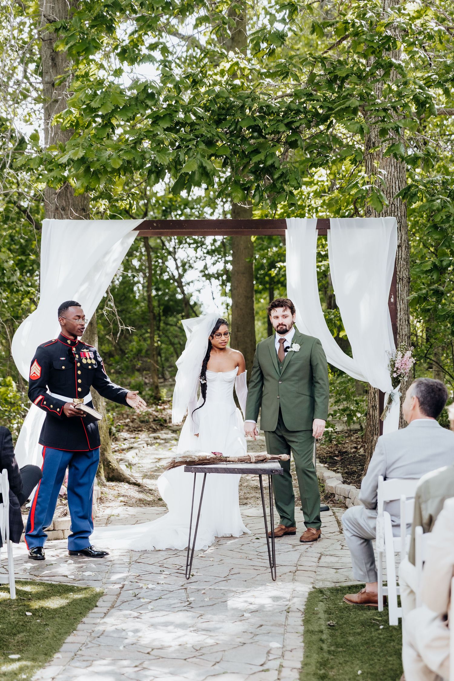 Wedding ceremony setup with white chairs and flowing fabric drapes between trees on a stone pathway.