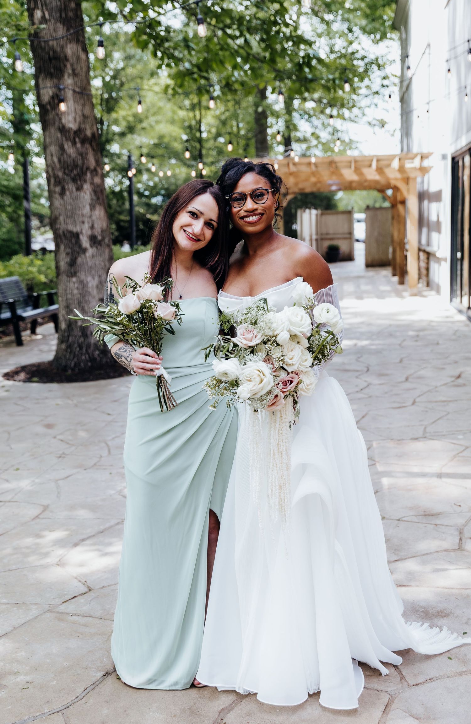 Two wedding attendees pose together holding beautiful white floral bouquets against stone wall backdrop.