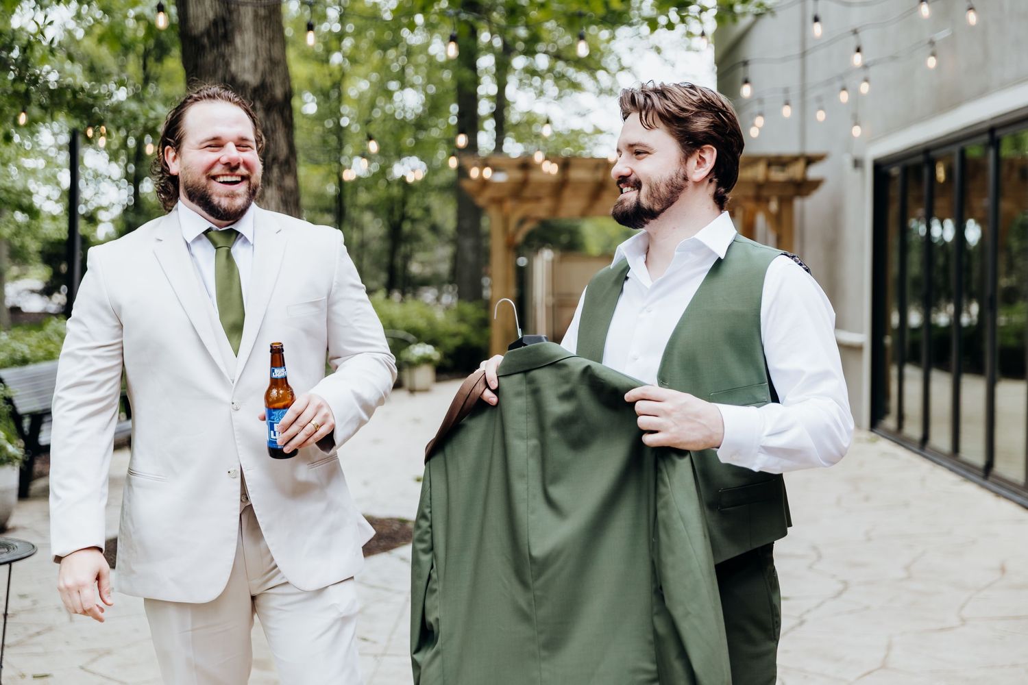 Two groomsmen share a laugh while holding drinks and a green suit jacket at an outdoor wedding venue.