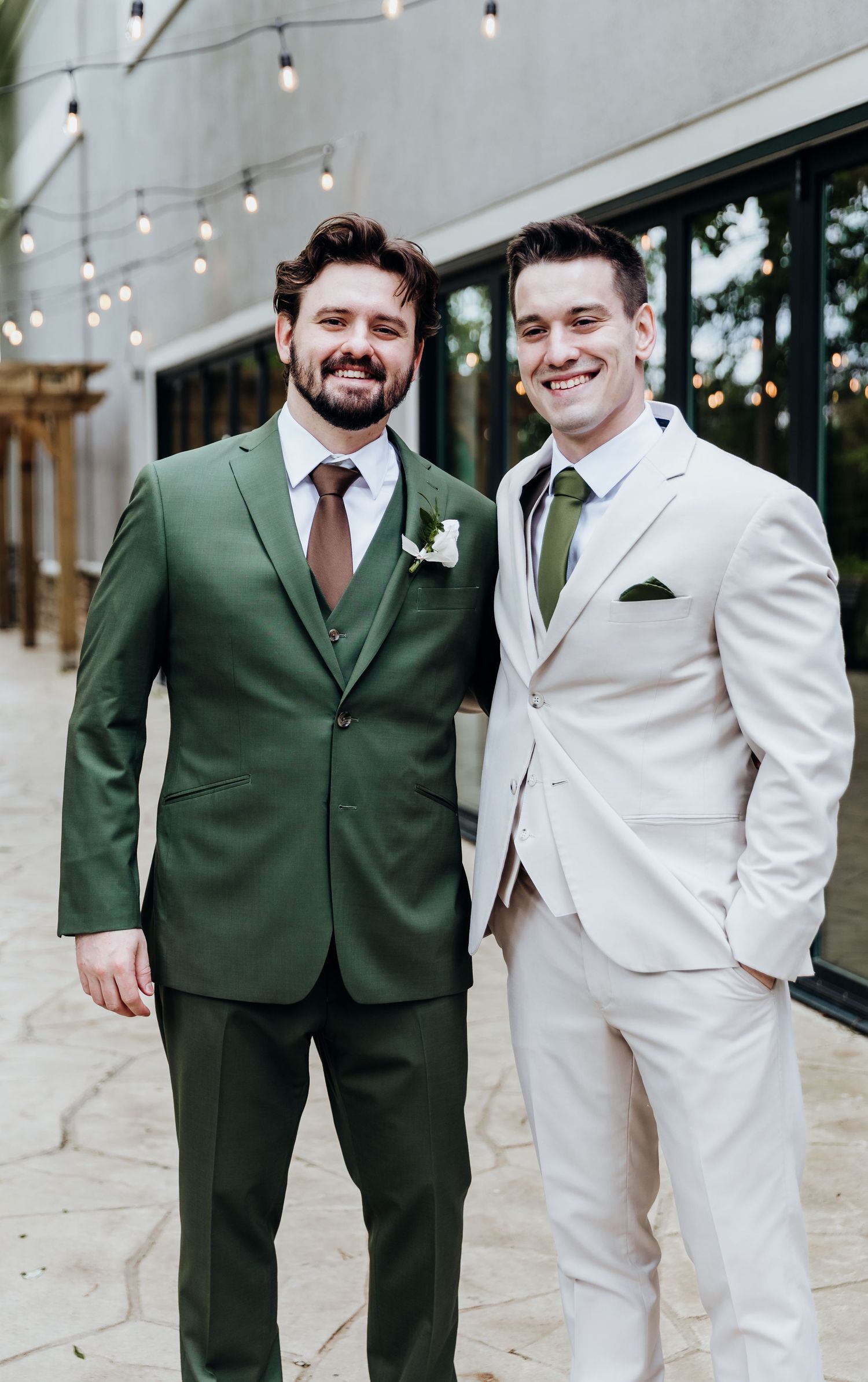 Two wedding attendants pose together wearing contrasting green and white suits with string lights in background.