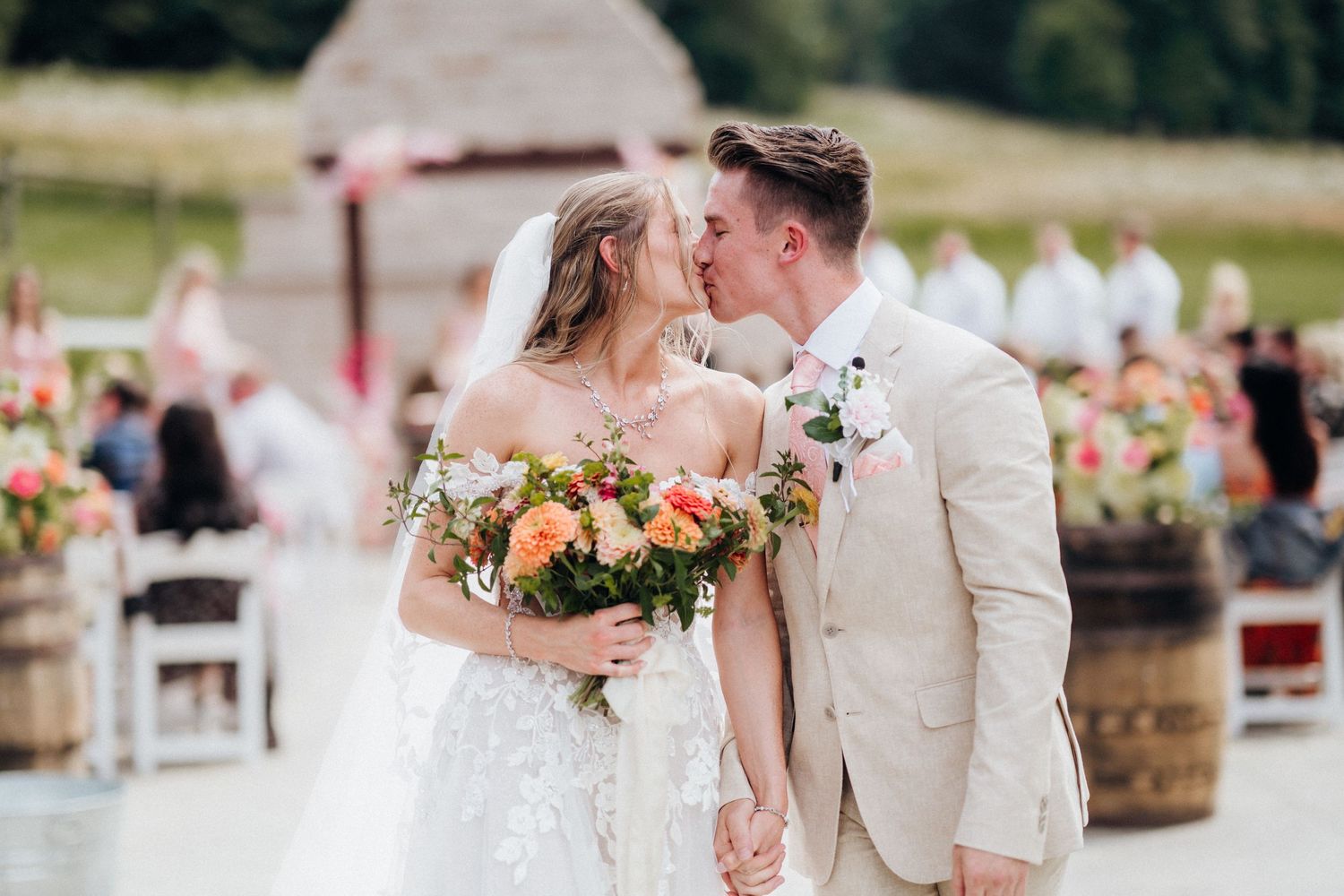 A romantic outdoor wedding ceremony with peach floral arrangements and rustic barrels.