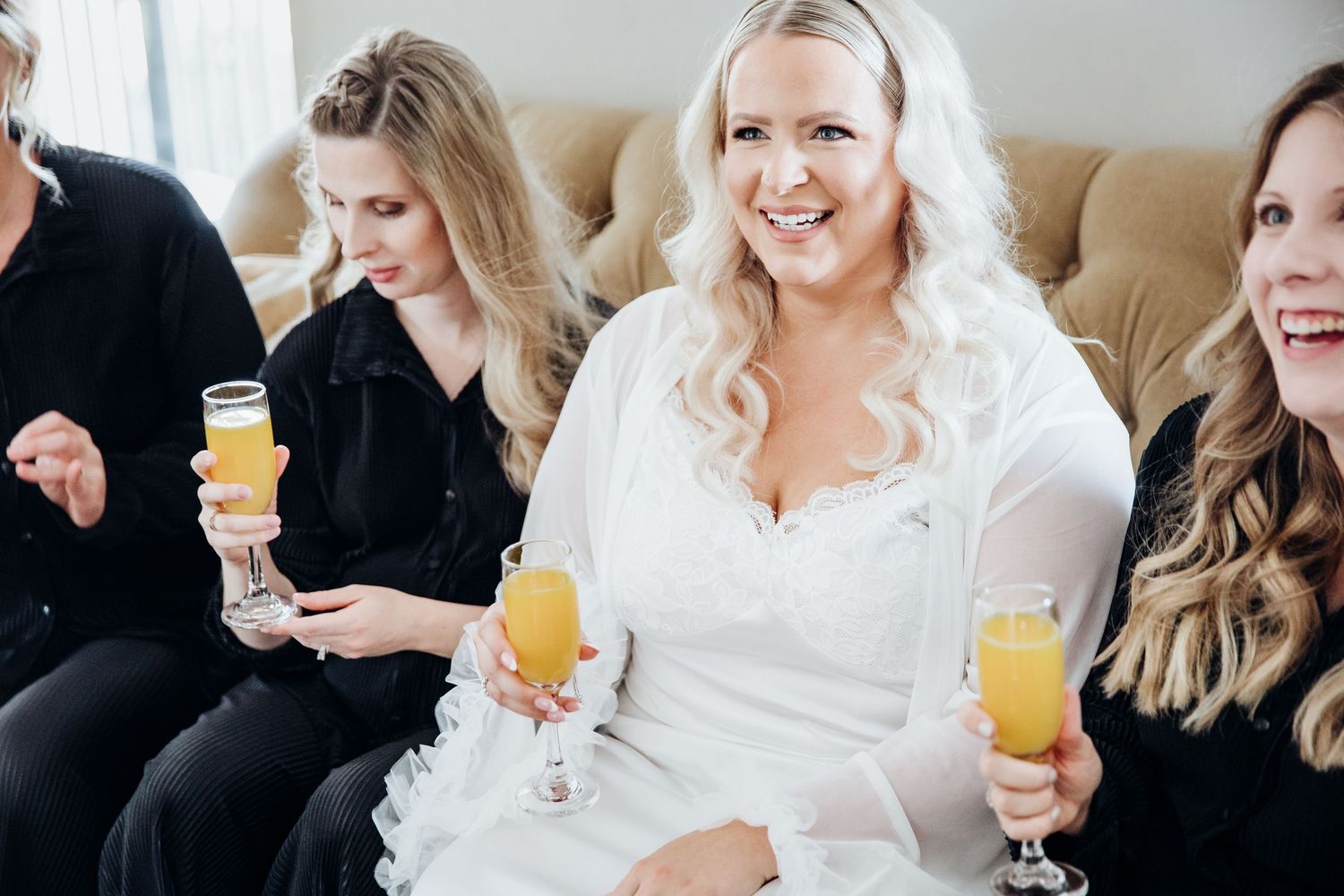Wedding party relaxes on beige couch with mimosas during pre-ceremony preparations.