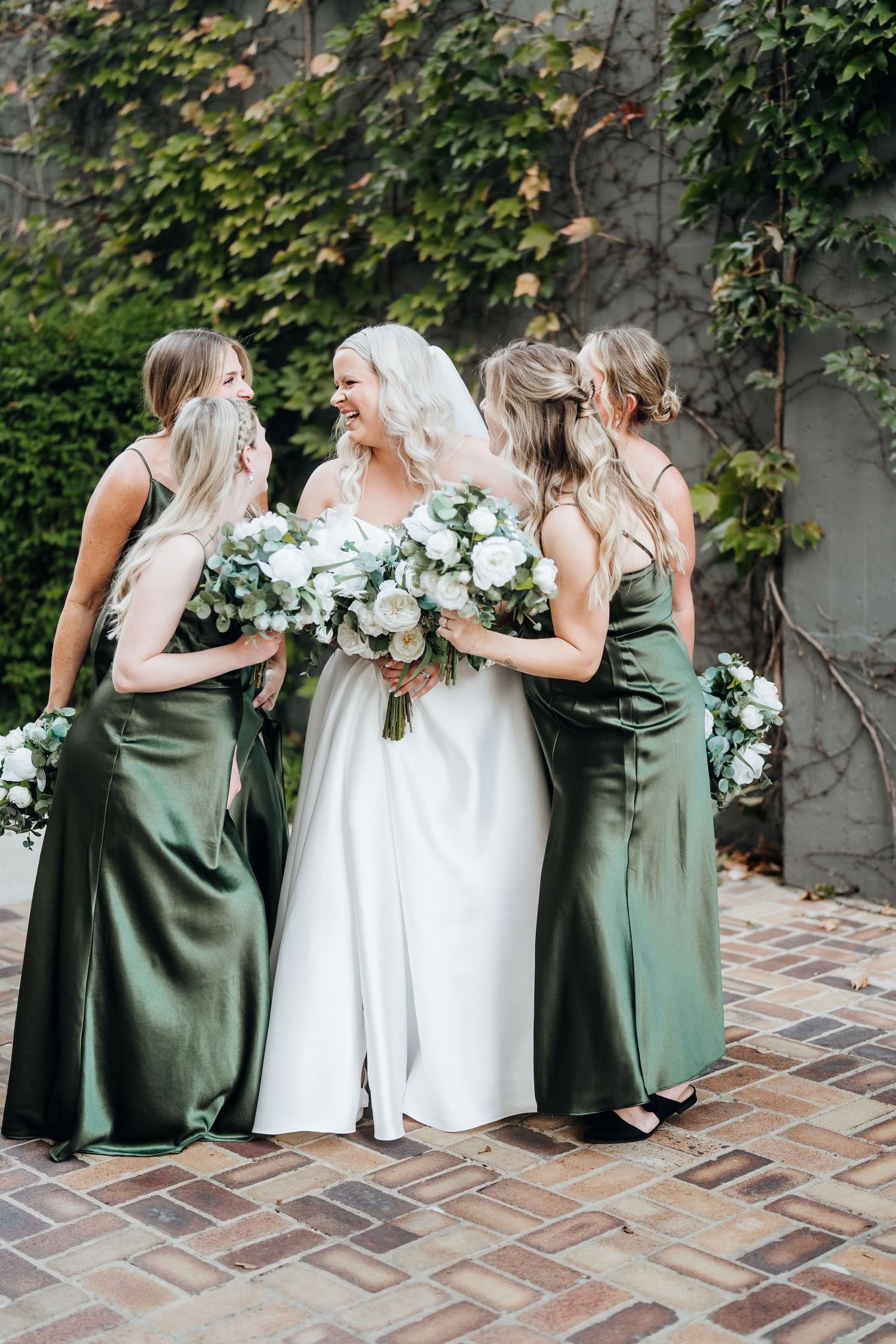 Bridesmaids in sage green dresses pose playfully with bride in white gown against ivy-covered wall.