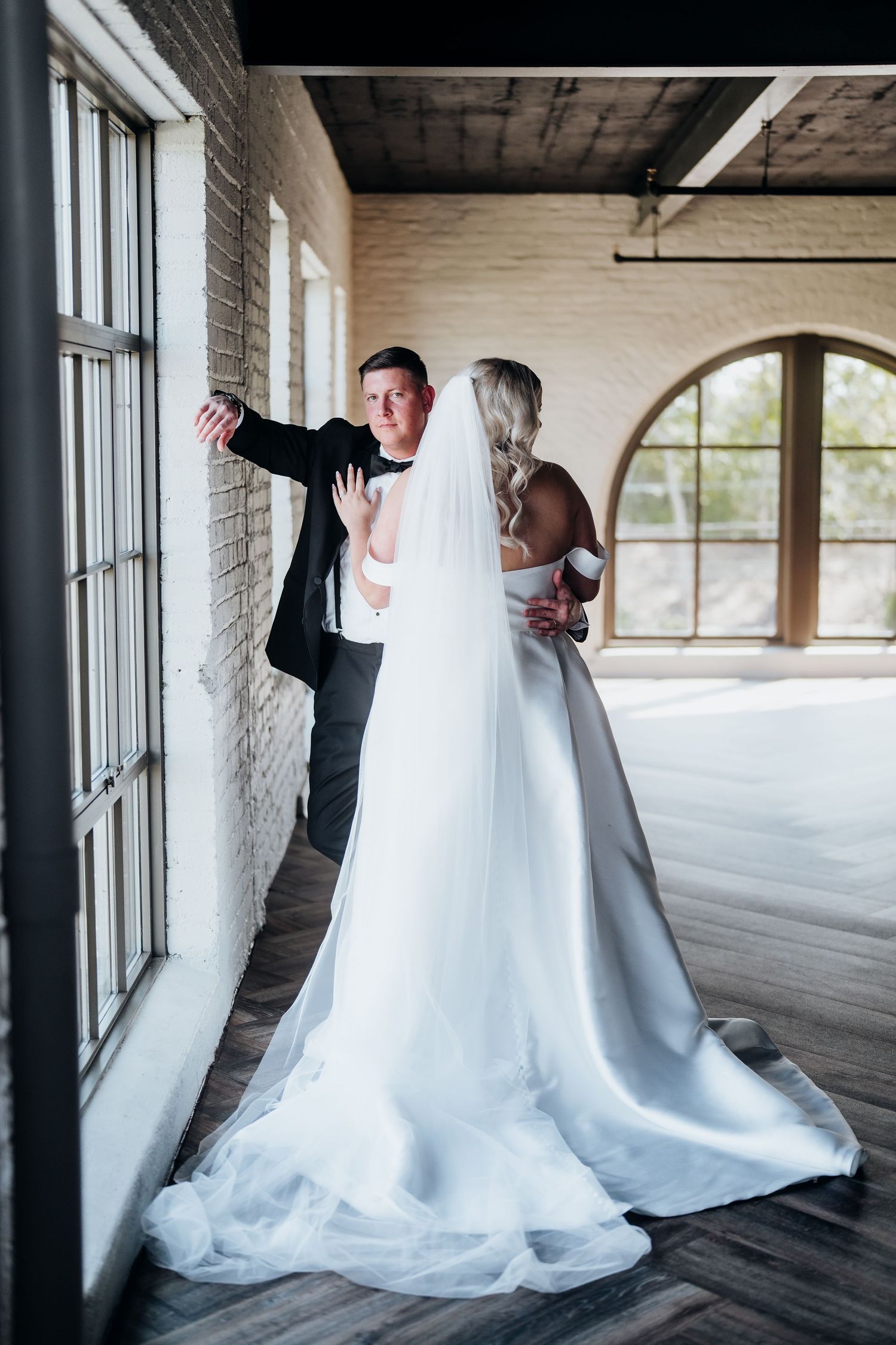 Wedding couple shares a romantic moment in front of large industrial windows.