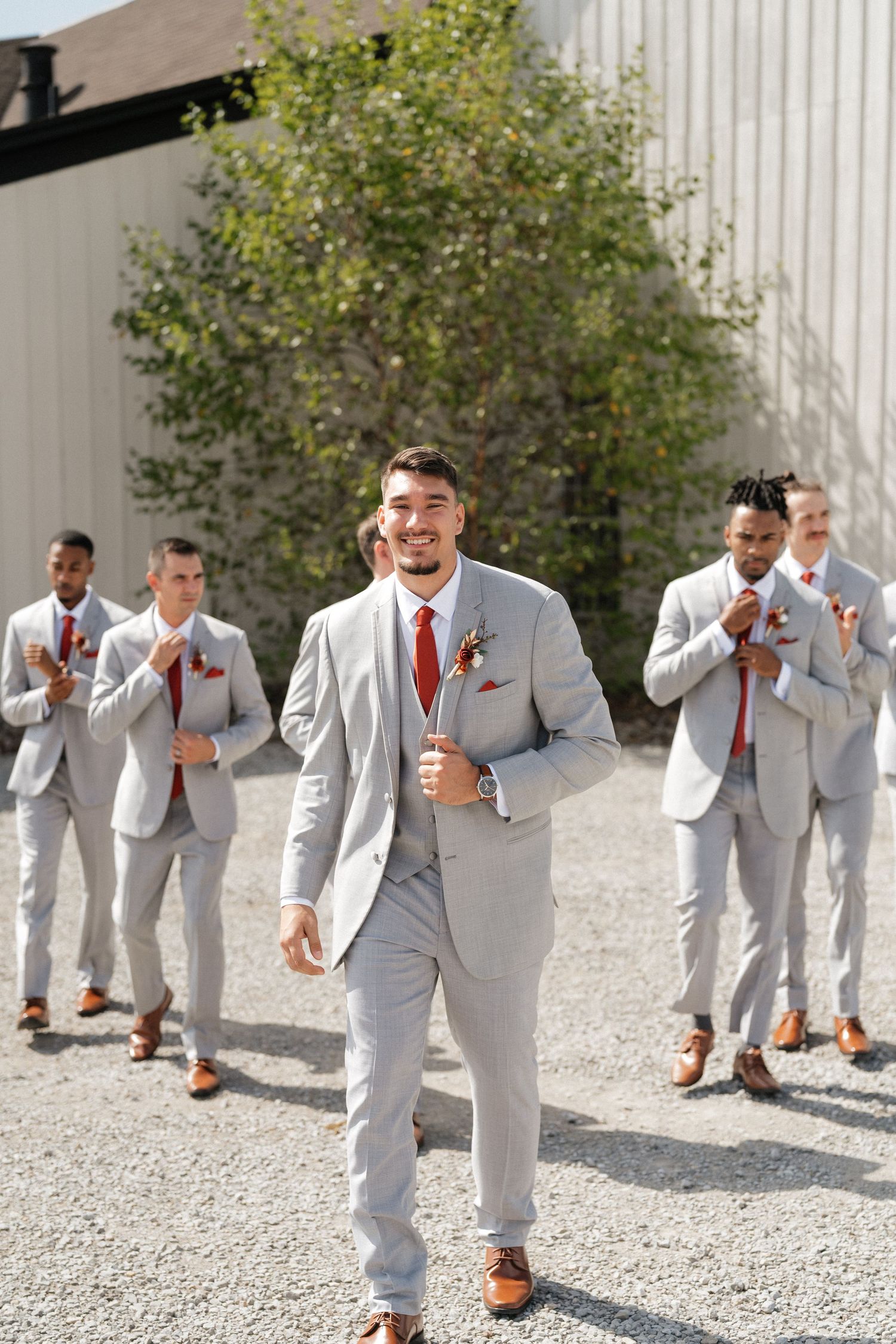 Groomsmen in light gray suits and red ties walk playfully on gravel path during wedding photos.