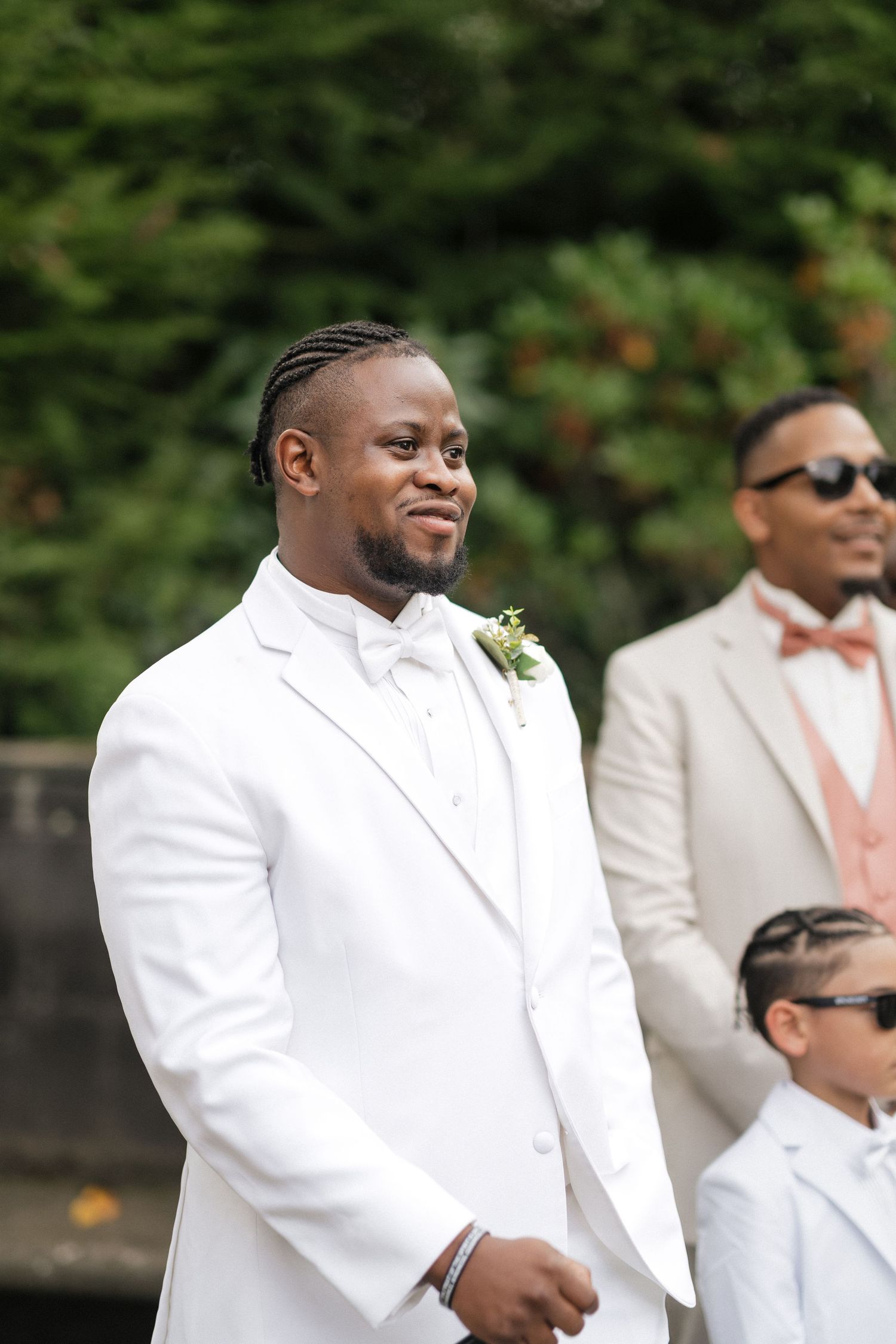Groomsmen in crisp white tuxedos and peach accents standing together at an outdoor wedding.