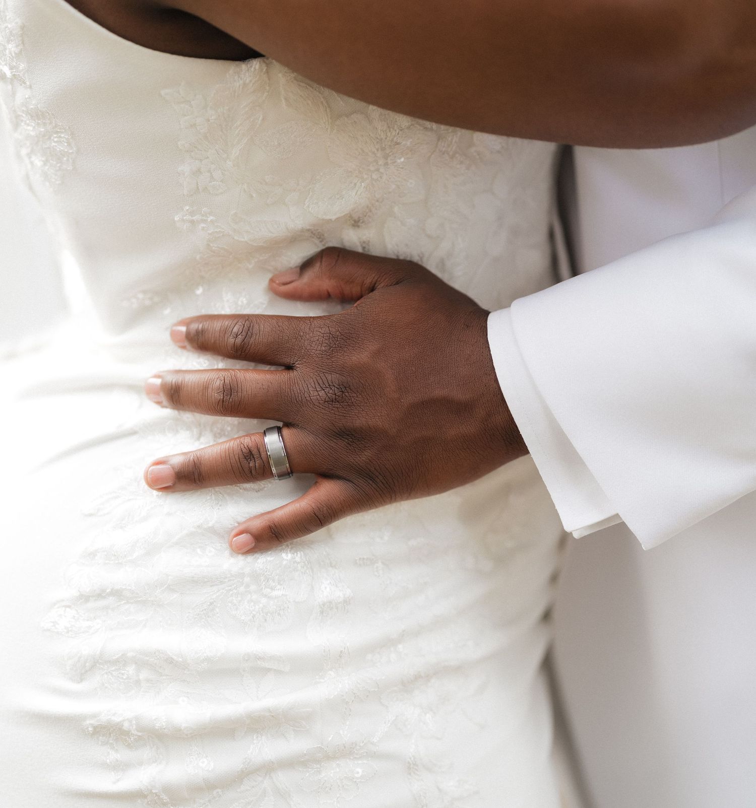 Close-up of hands with wedding ring against white dress showing tender embrace during wedding celebration.