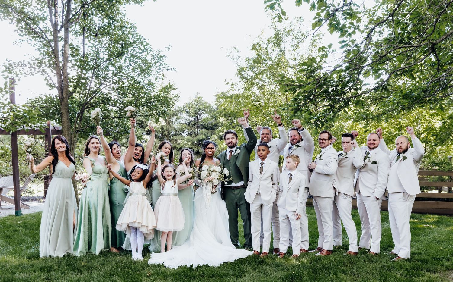 Large wedding party in white and mint green attire poses for group photos in garden setting.