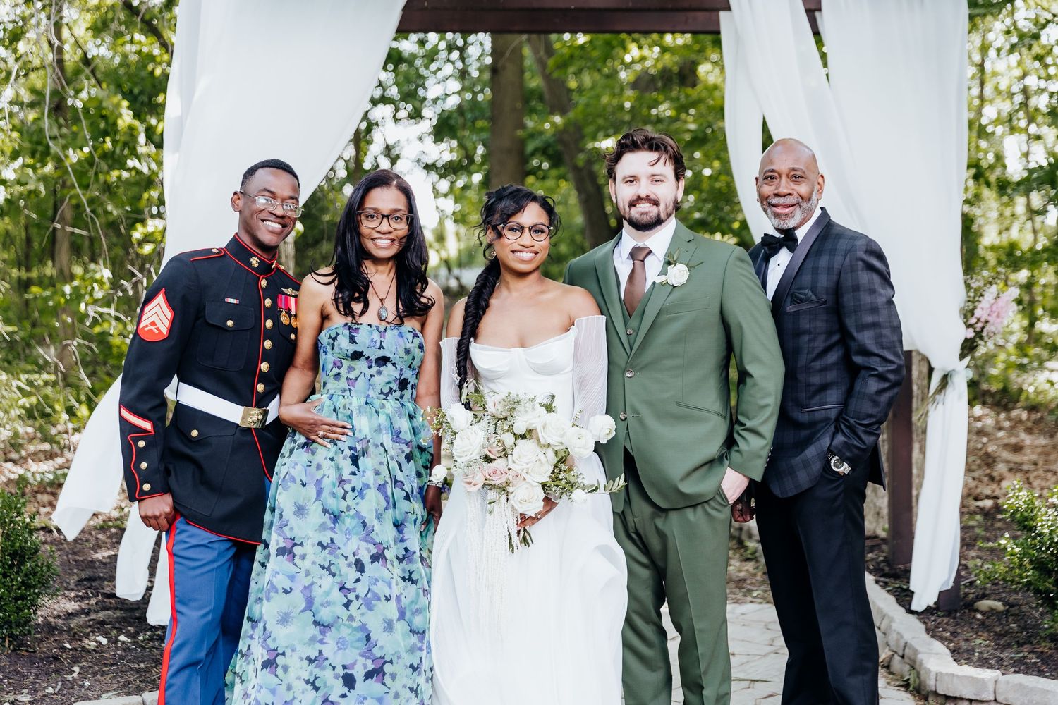 Wedding party poses together under white arch with mix of formal attire in green and blue tones.