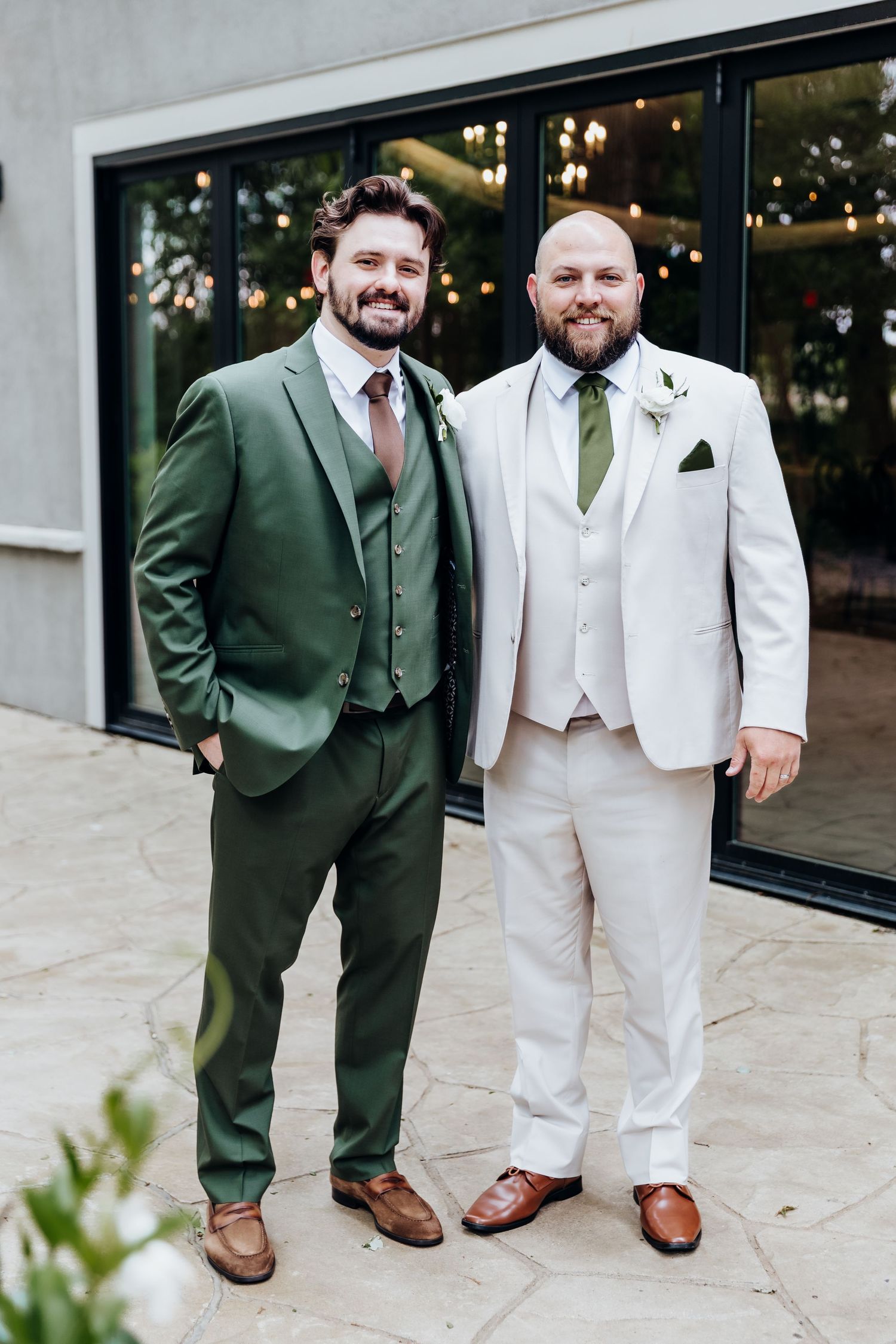 Two people in contrasting green and white suits stand together showing off their wedding attire and brown dress shoes.