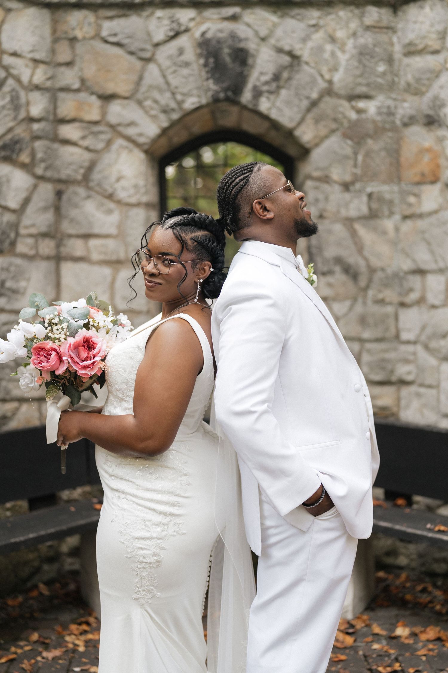 Wedding couple shares a playful moment in front of a stone wall while holding a pink rose bouquet.
