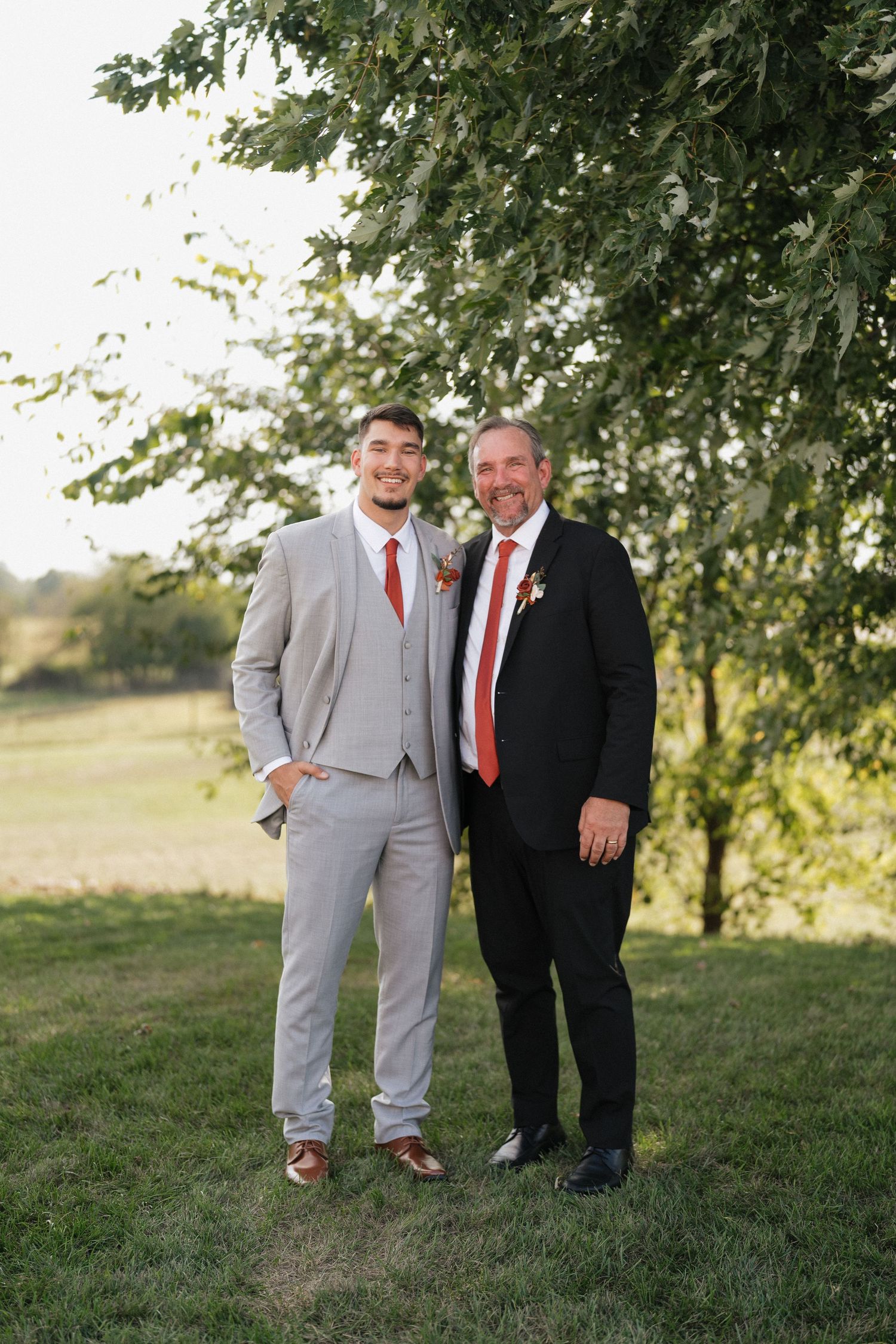 Wedding party members in formal attire posing under trees during outdoor photo session.