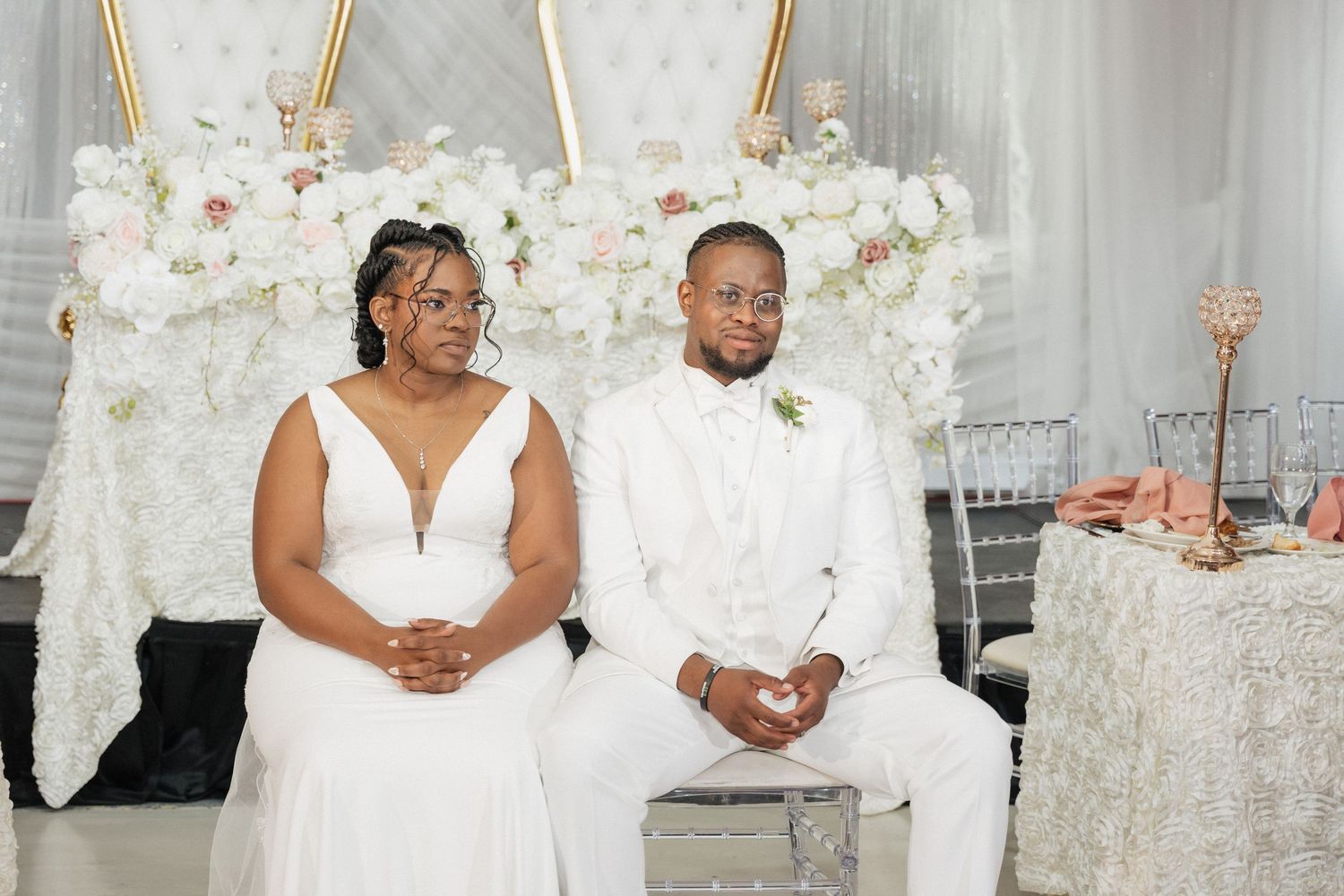 A couple sits together at their wedding reception with white floral arch and decor.