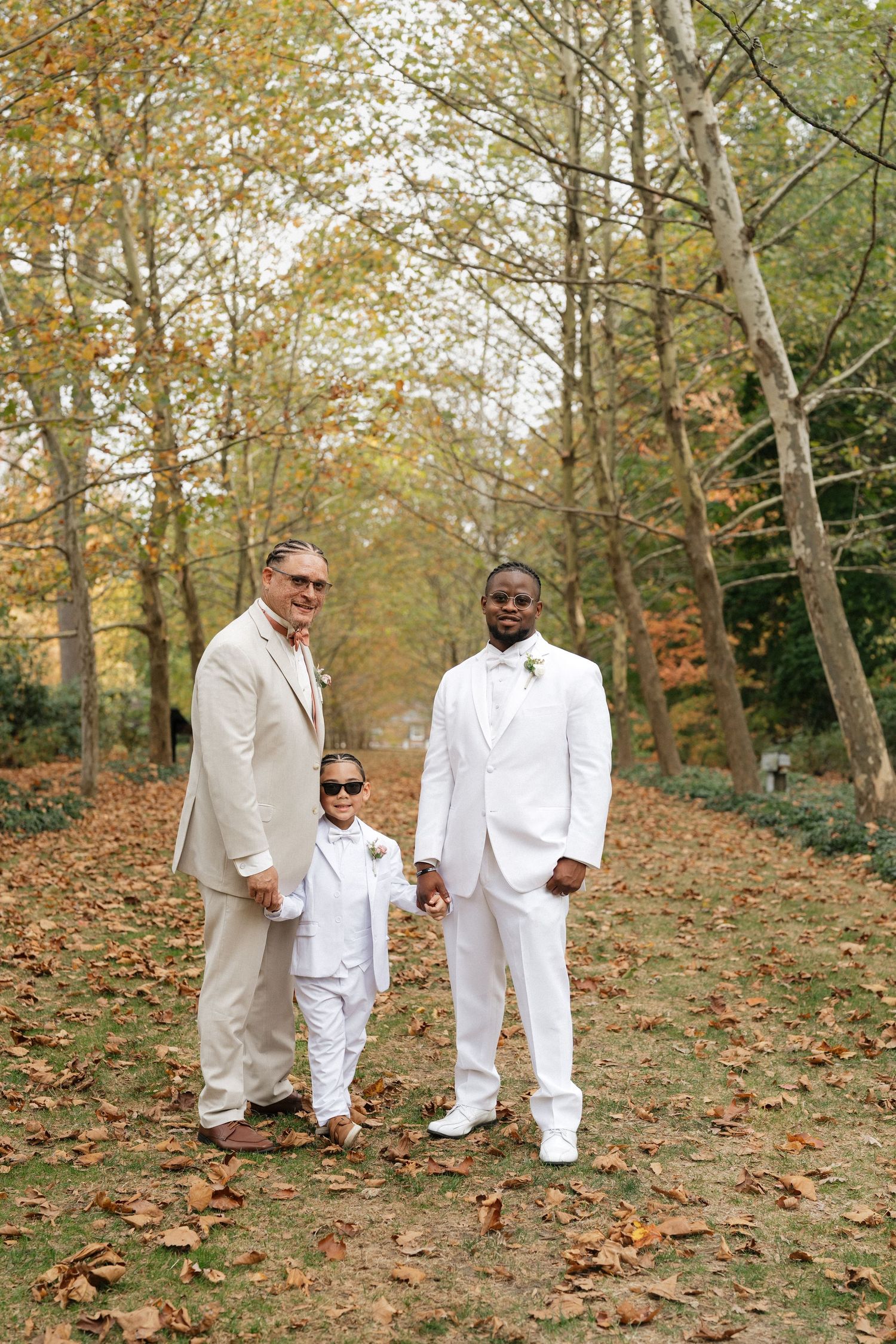 Three people in coordinating white and beige suits pose on a leaf-covered forest path.
