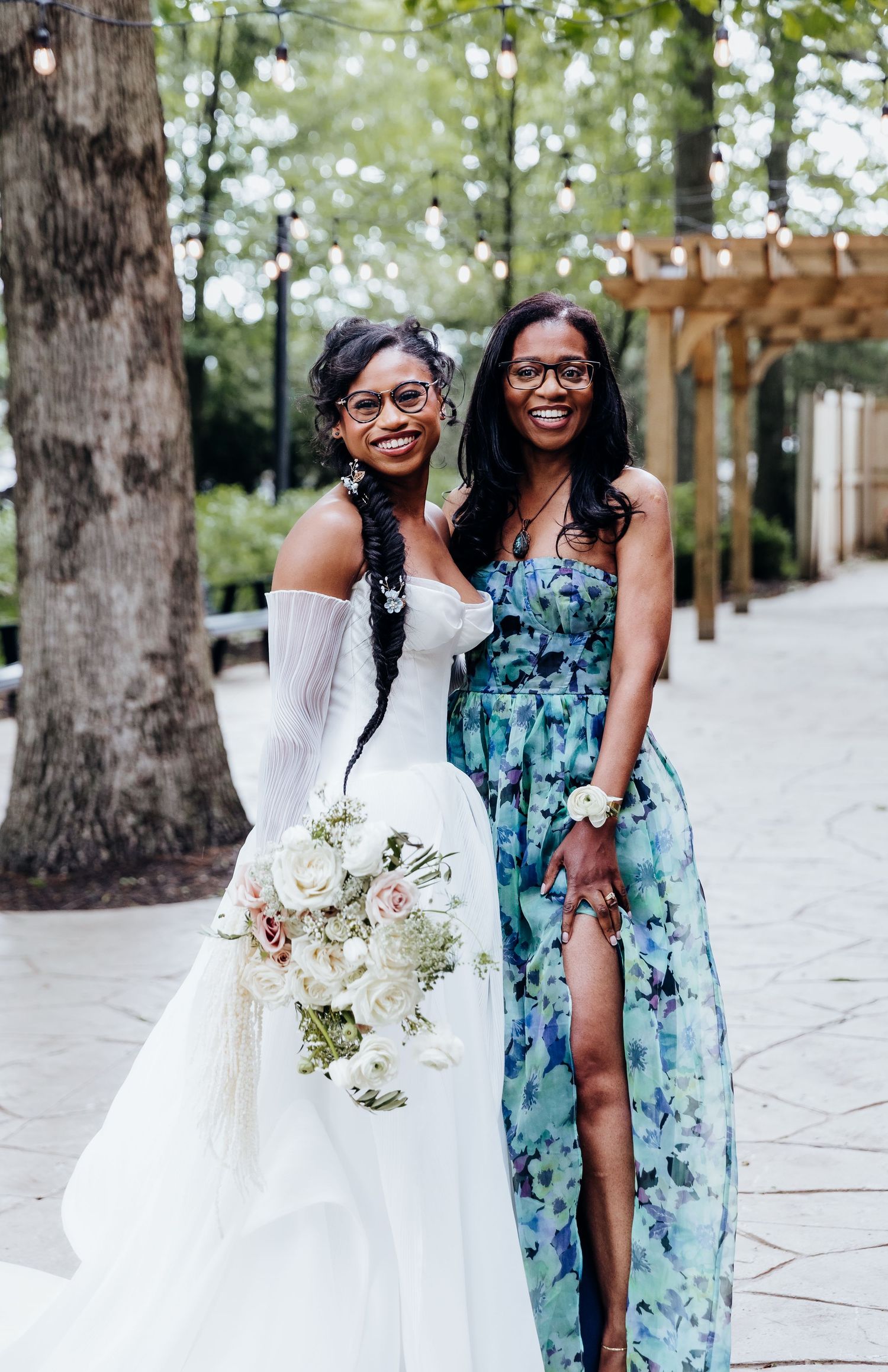 Two wedding guests in elegant dresses pose together outdoors near trees.