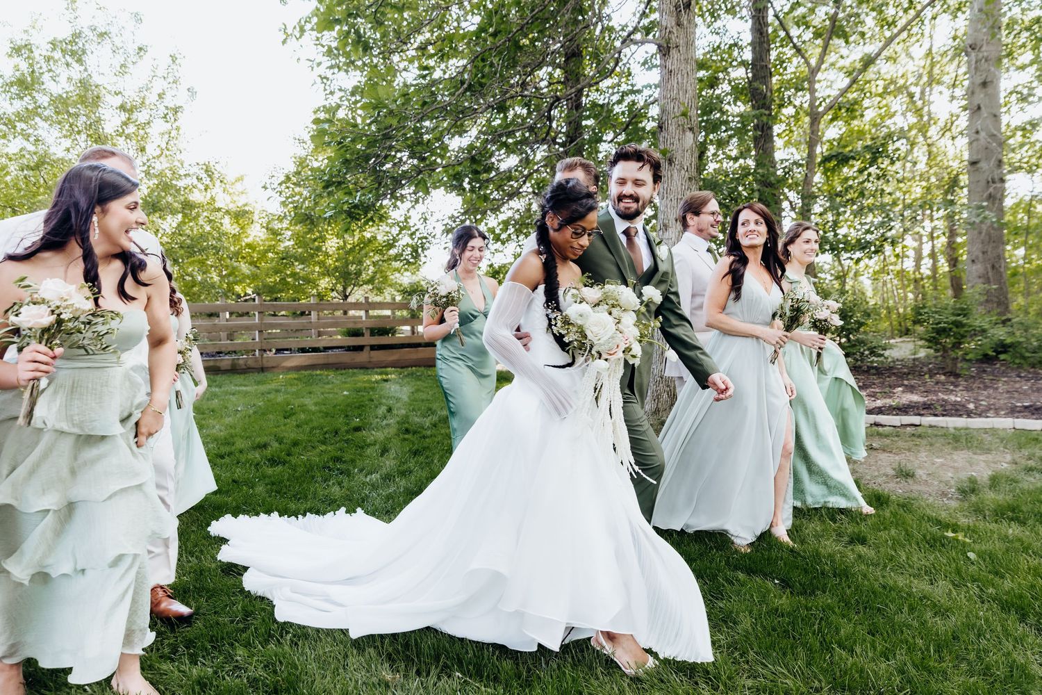 Wedding party members in mint green dresses run playfully through grassy outdoor setting.