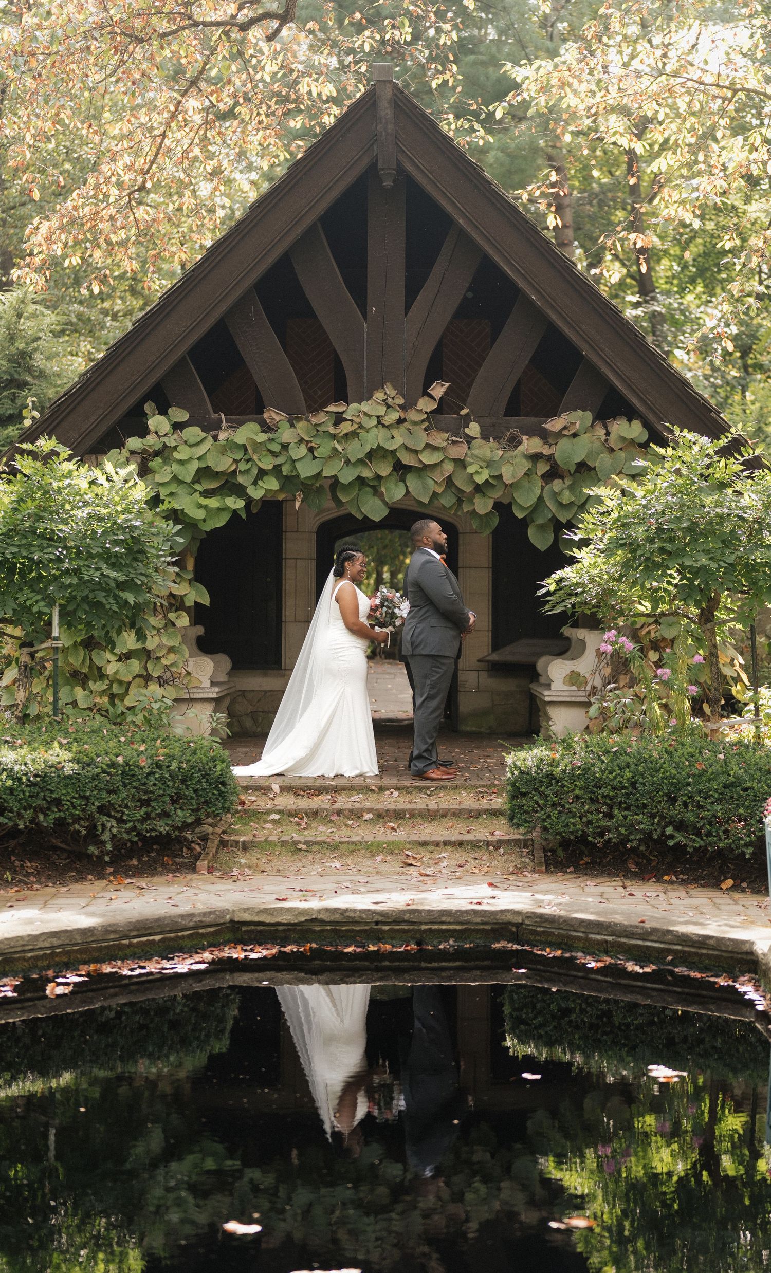 A wedding couple stands in a rustic wooden gazebo with reflecting pool and blooming flowers.