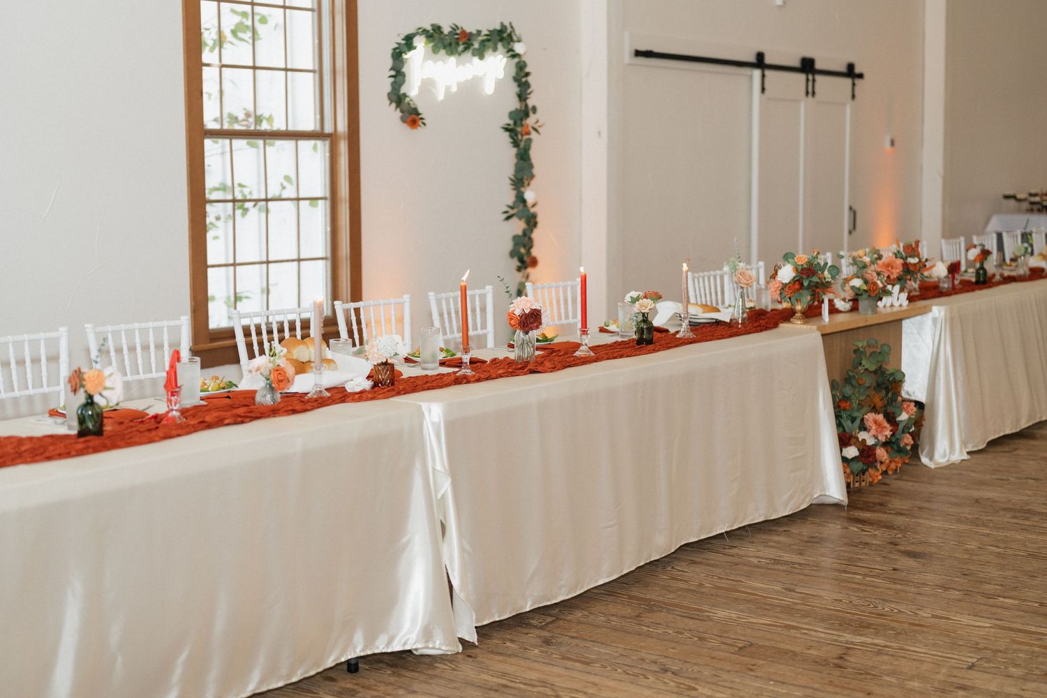 Long white reception table with orange accents and greenery garland decor.