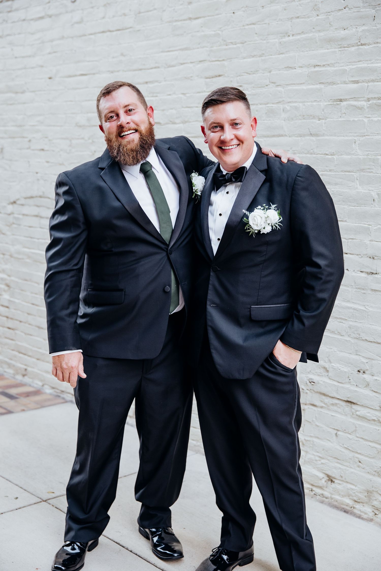 Two groomsmen in black tuxedos pose against a white brick wall.